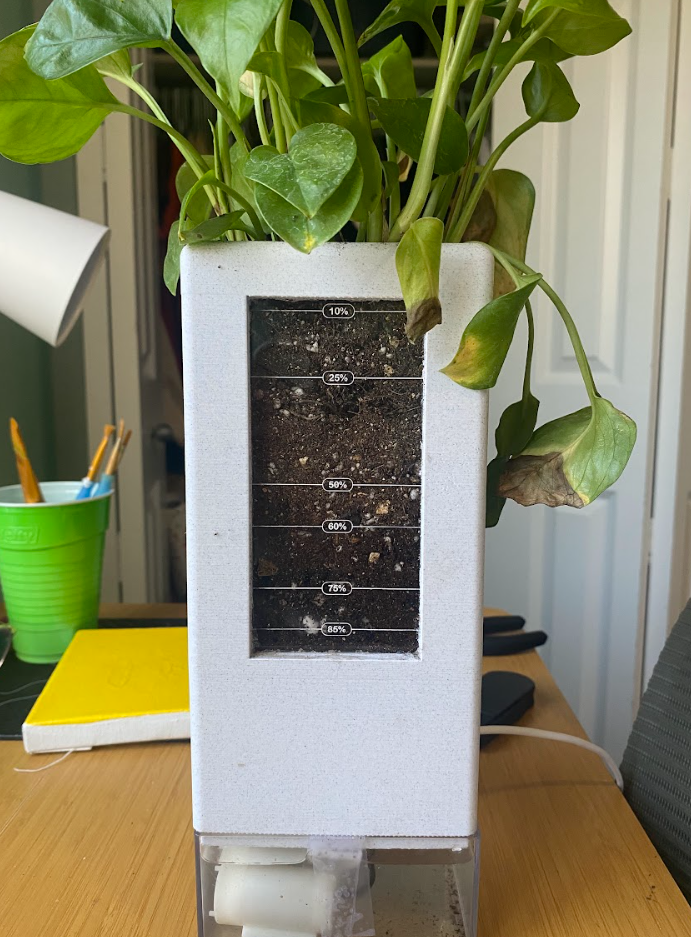 White self-watering planter on a desk with a leafy pothos, its soil-level window marked in percentages.