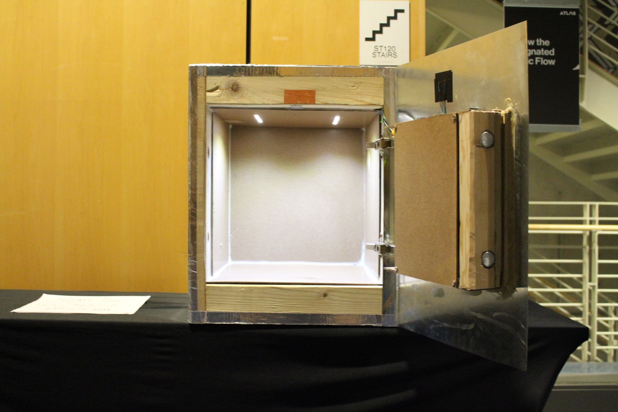 A metal-framed wooden box with its hinged door open, white-lined interior lit by inner LEDs, on a display table.