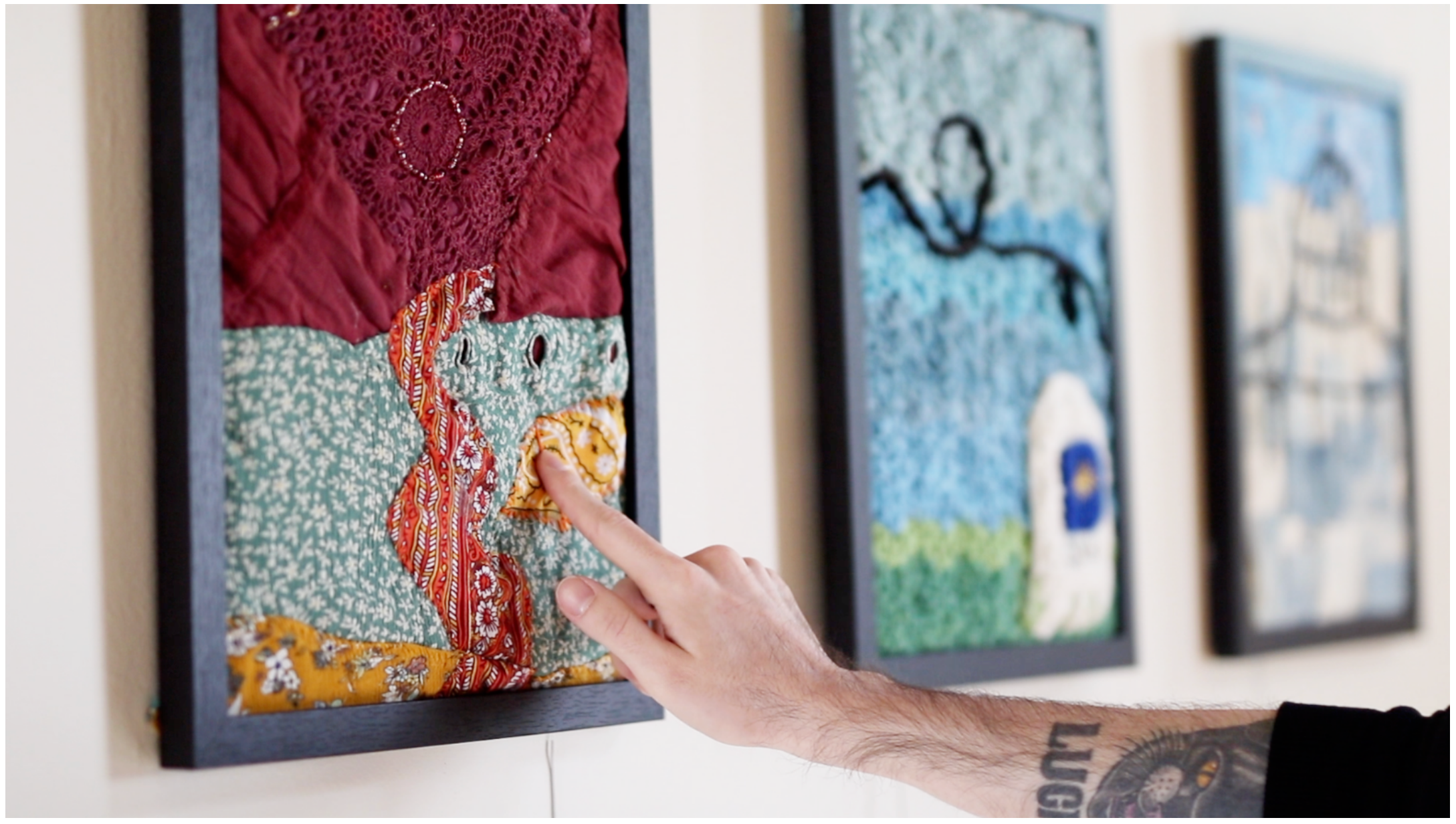 A finger touches a framed fabric collage on a gallery wall; two more textile panels hang alongside.