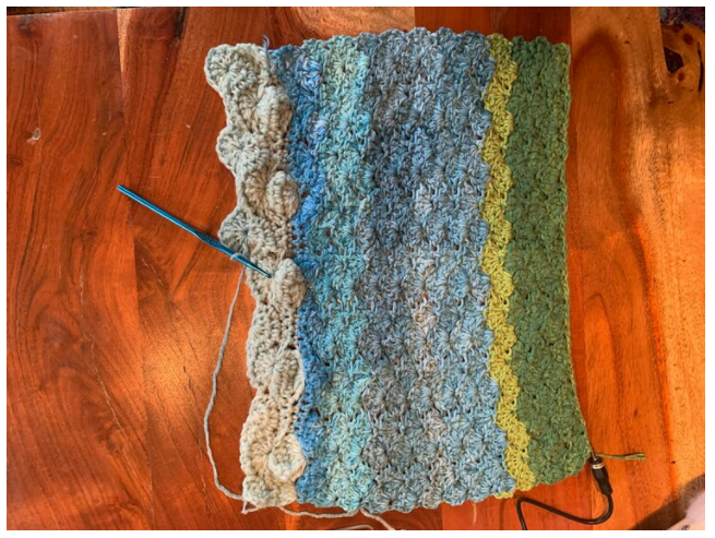 A crocheted panel in beige, blue, and green stripes with a hook resting on a wood table.
