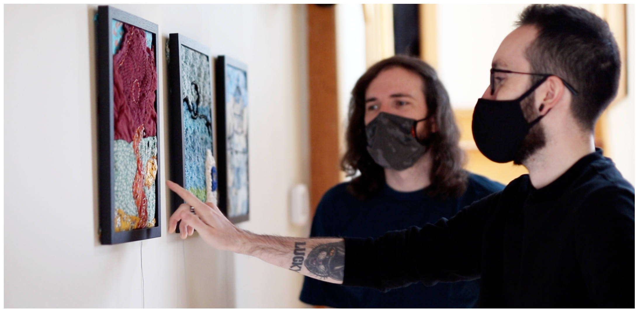 Two masked men view framed textile artworks on a gallery wall; one points at a touch-sensitive panel.
