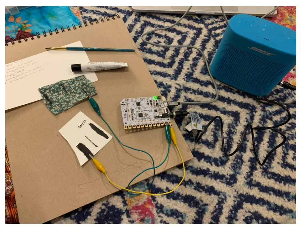 A cardboard workbench with a microcontroller wired by clip leads to fabric swatches beside a Bose speaker.