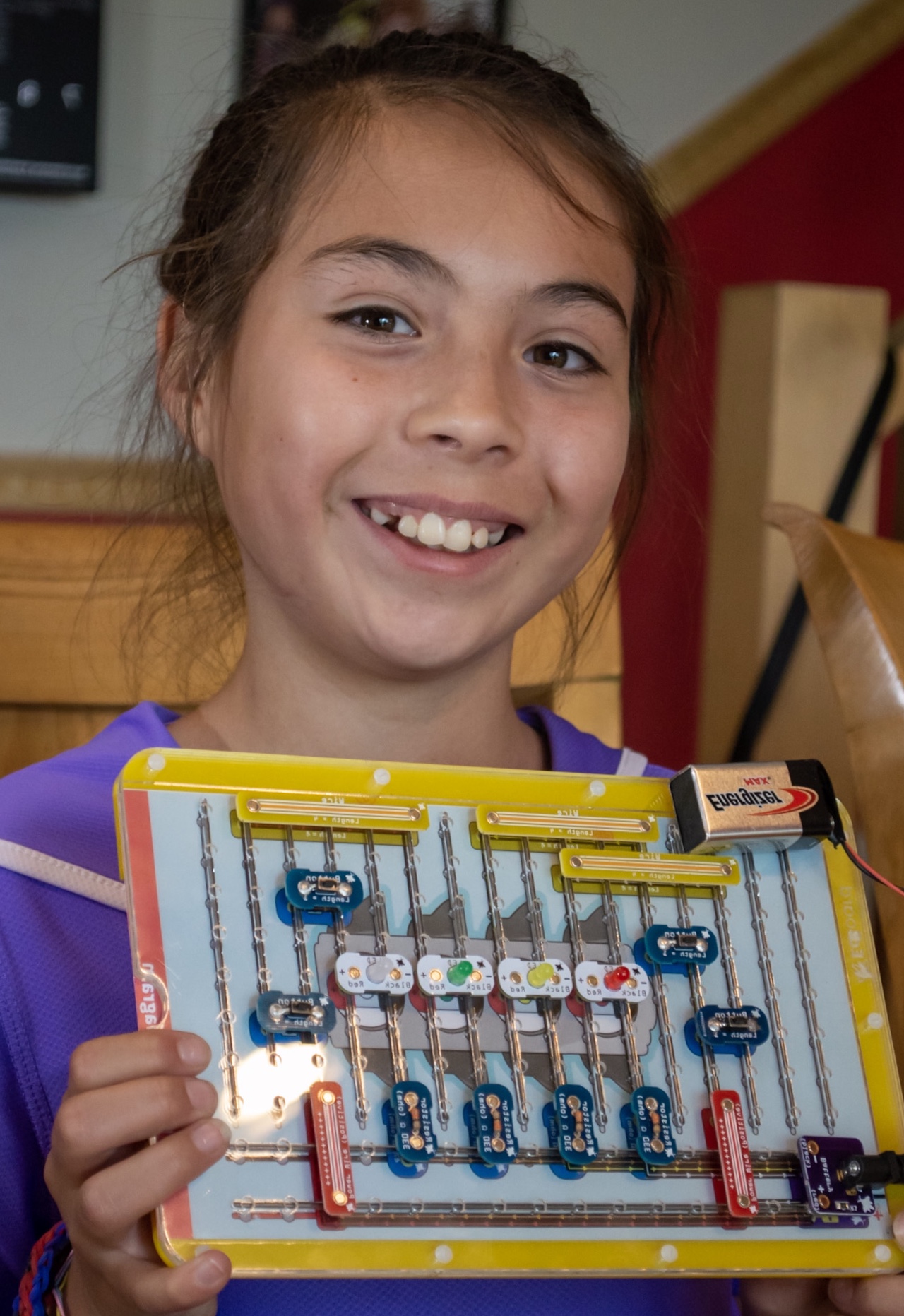 A smiling girl holds up a yellow circuit-building board strung with lit LEDs and a 9-volt battery.