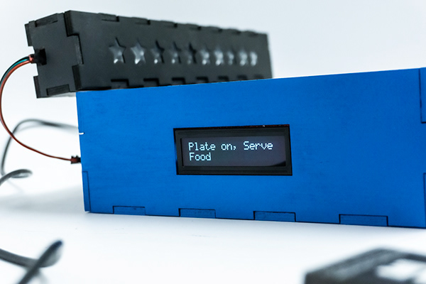 Close-up of the blue plate device, LCD reading "Plate on, Serve Food" beside a star reward bar.