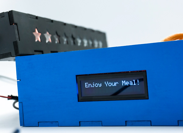 Close-up of the blue plate device with an LCD reading "Enjoy Your Meal!" and lit star icons.