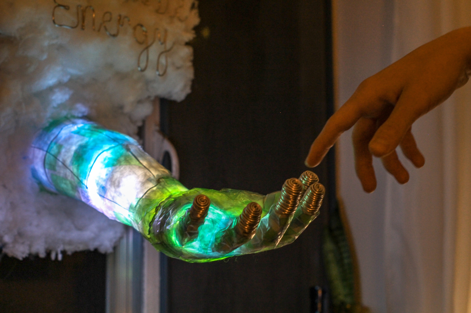 A glowing translucent sculpted arm and hand, lit green and blue, reaches toward a visitor's pointing finger.
