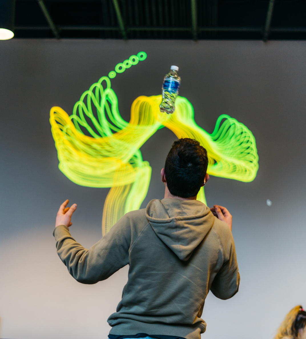 A person before a screen that traces their motion as glowing green ribbons around a floating water bottle.