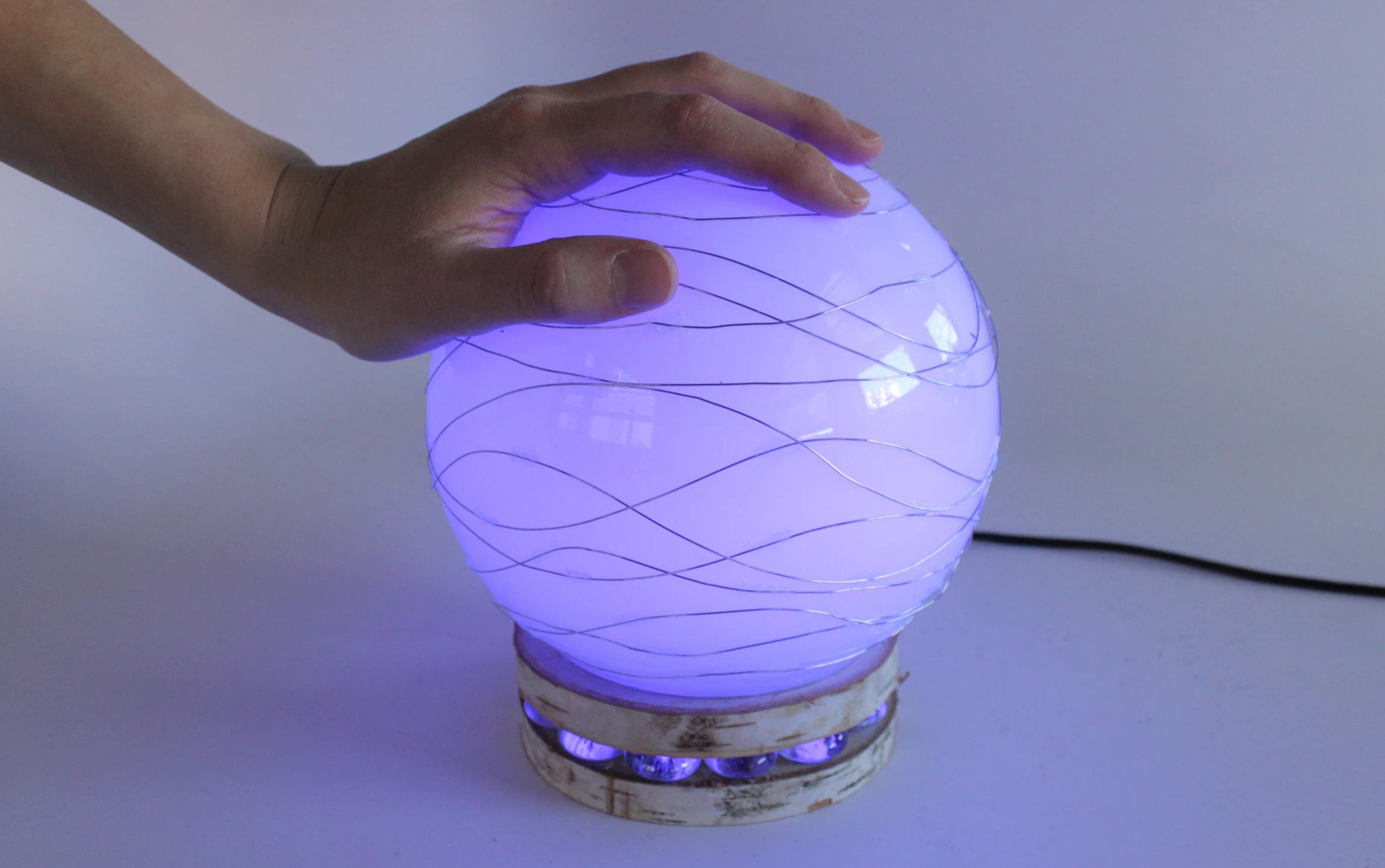 A hand rests on a glowing purple orb wrapped in wavy wires, set on a small wooden base.