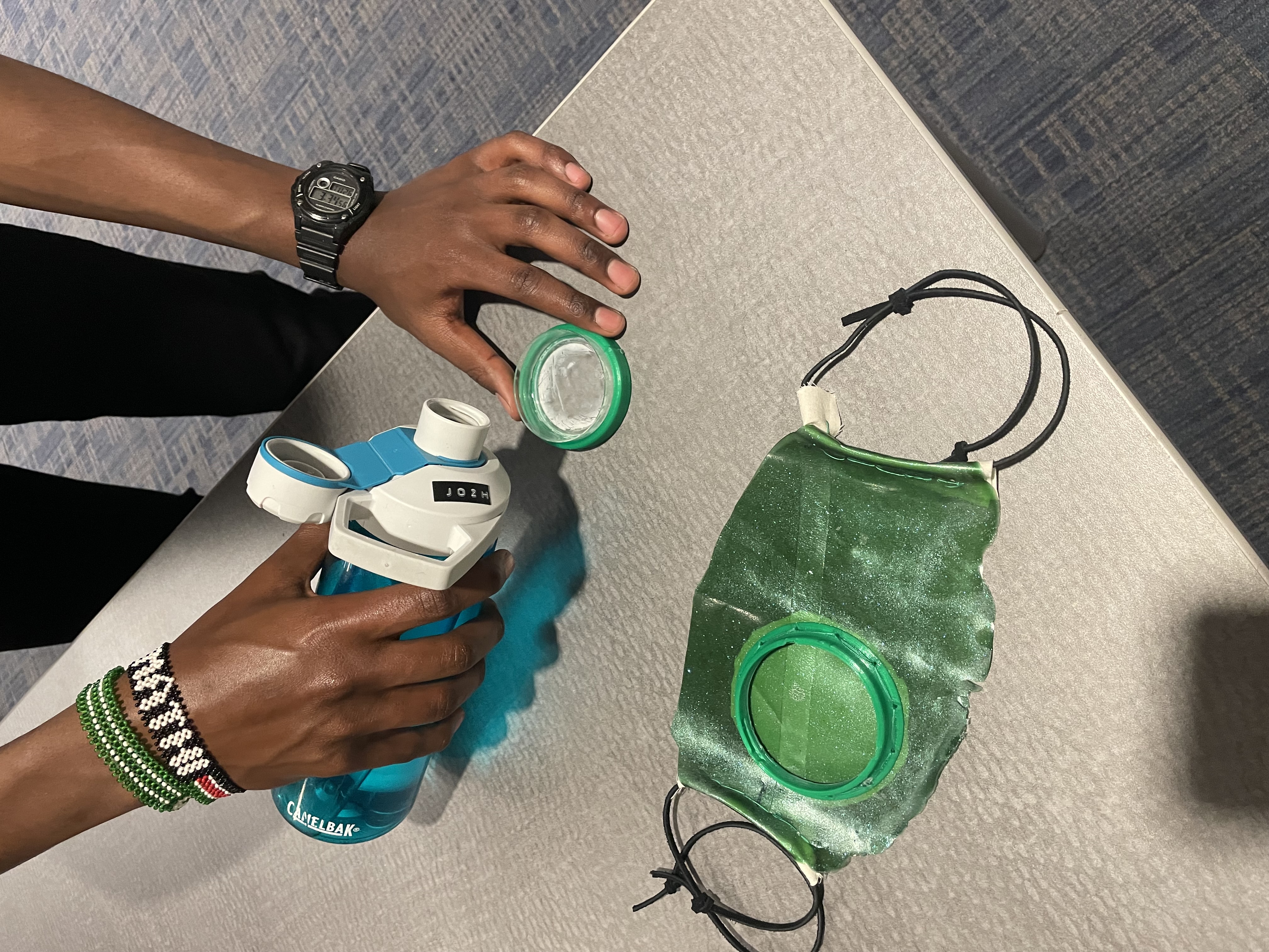 Hands hold the green Petri-dish mask beside a water bottle; the dish lid lifts off in one hand.