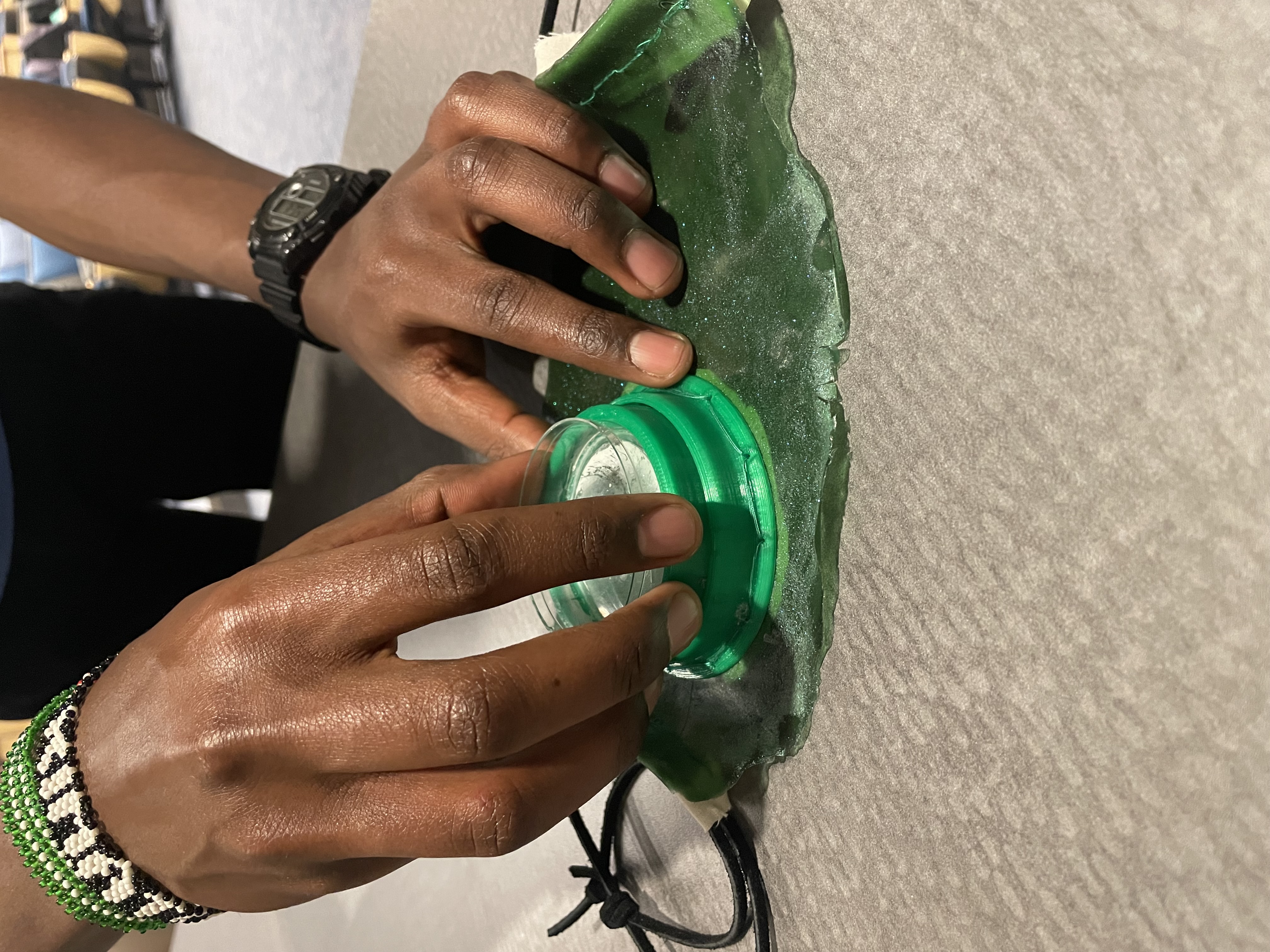 Hands press the round green Petri-dish module into the algae-textured fabric of the mask.