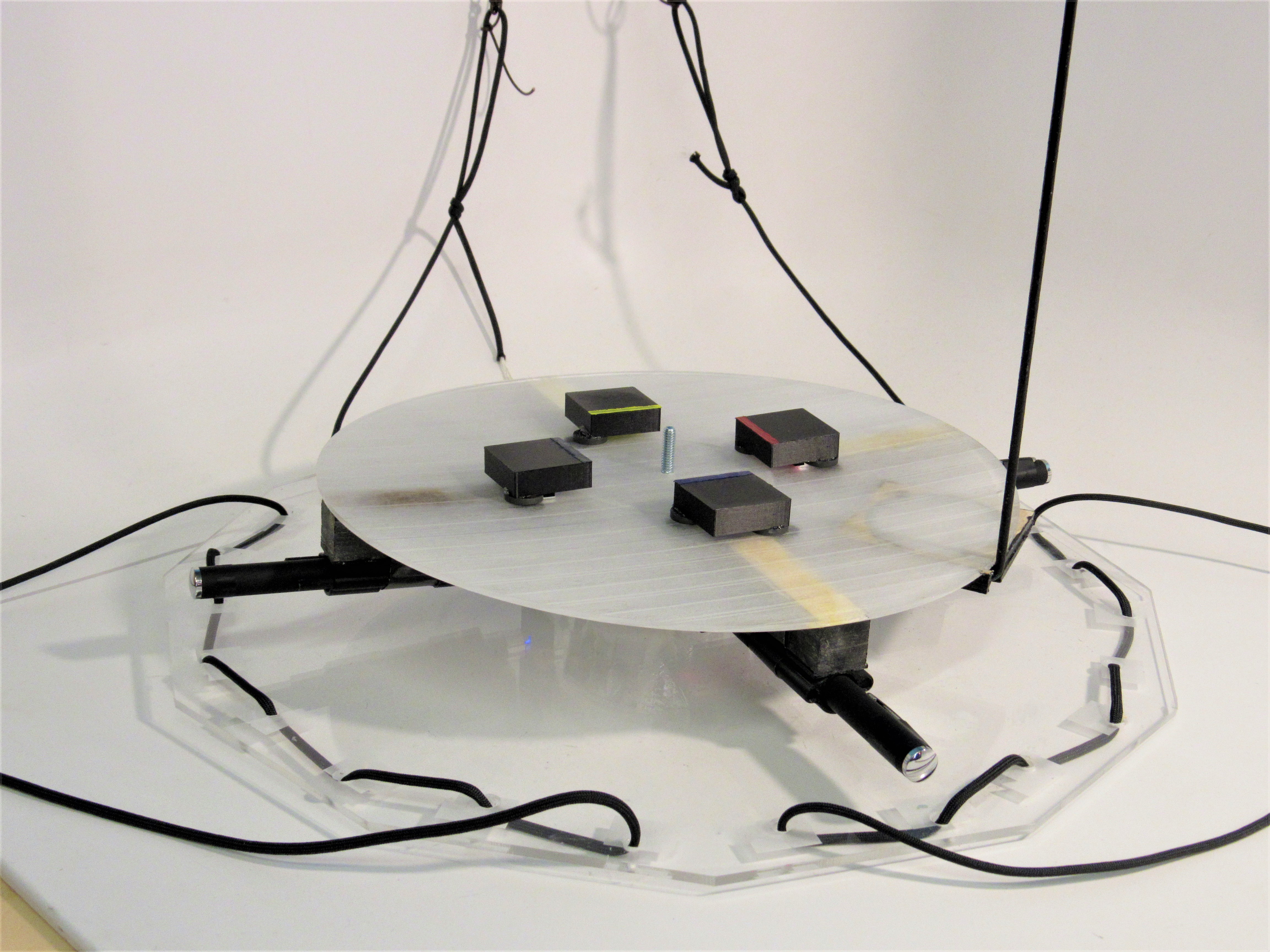 Suspended acrylic sculpture: a round metal plate holds four small color-coded blocks, wired up.