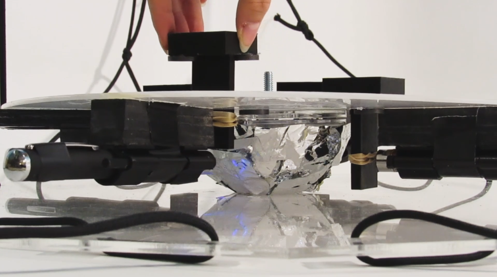 A fingertip presses a 3D-printed knob above the foil-and-laser center of the sculpture.