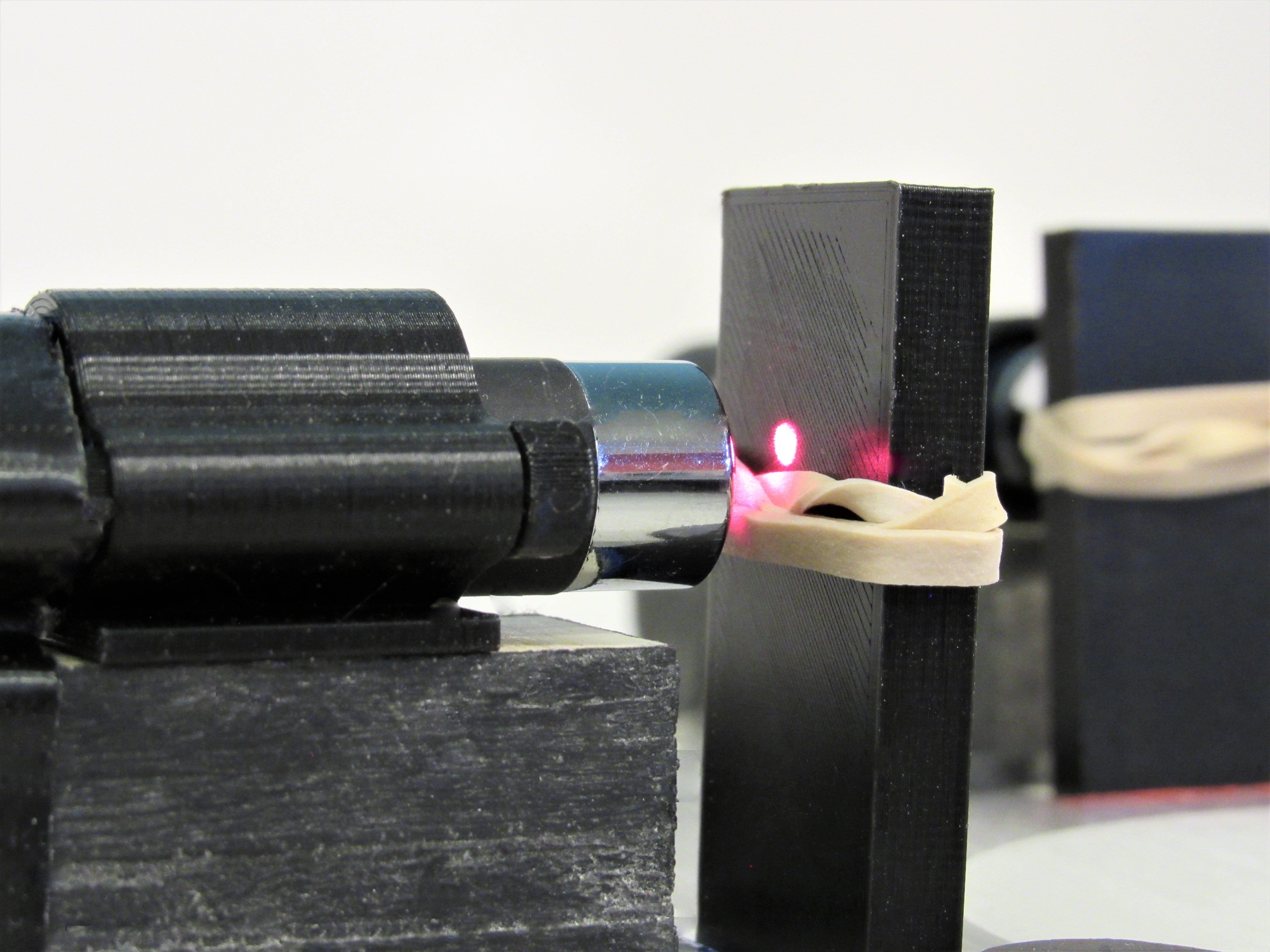 A chrome laser pointer fires a red beam at a rubber band stretched across a black post.