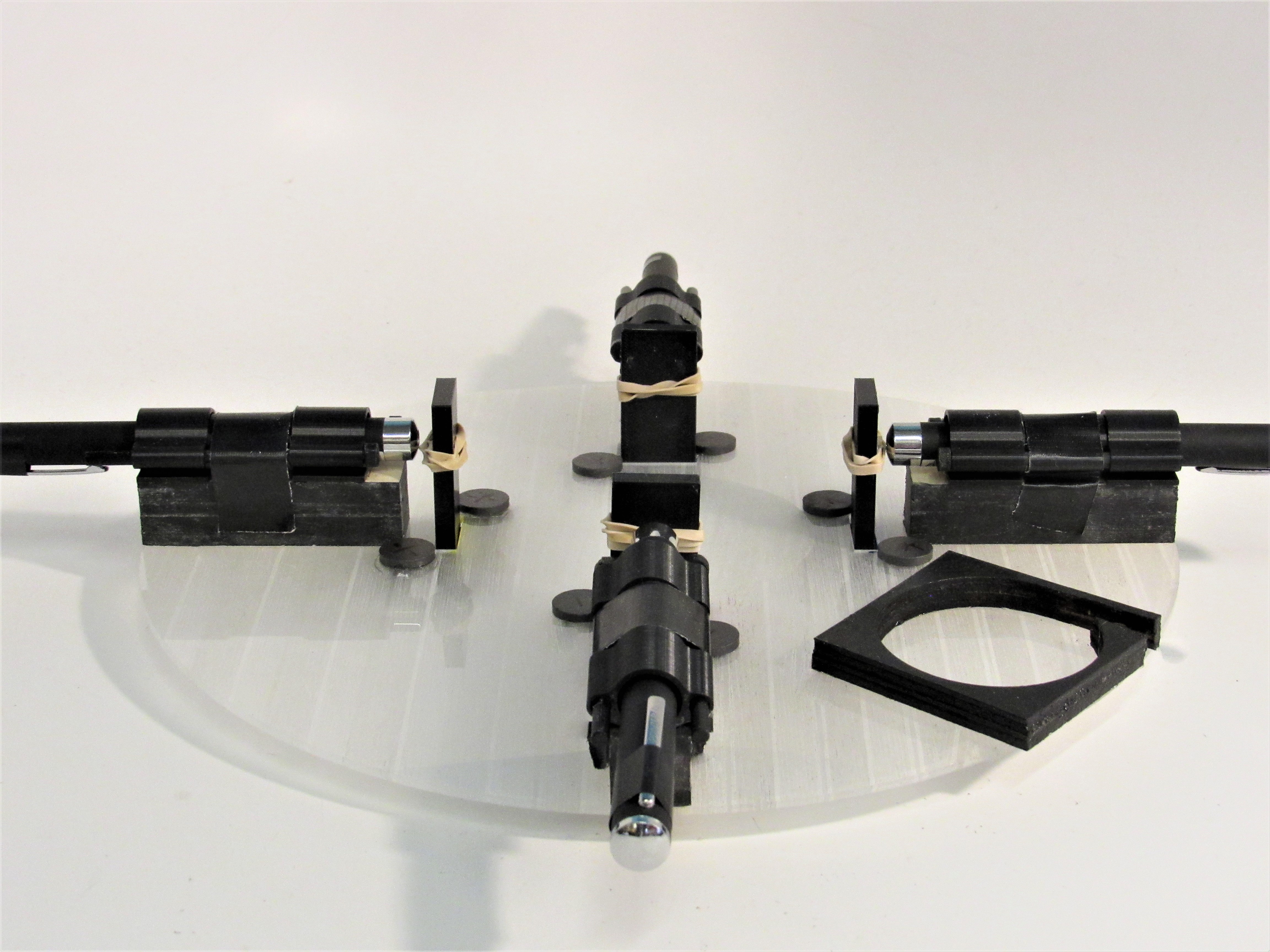 Disassembled 3D-printed laser mounts and chrome pointers laid out on a clear acrylic disc.