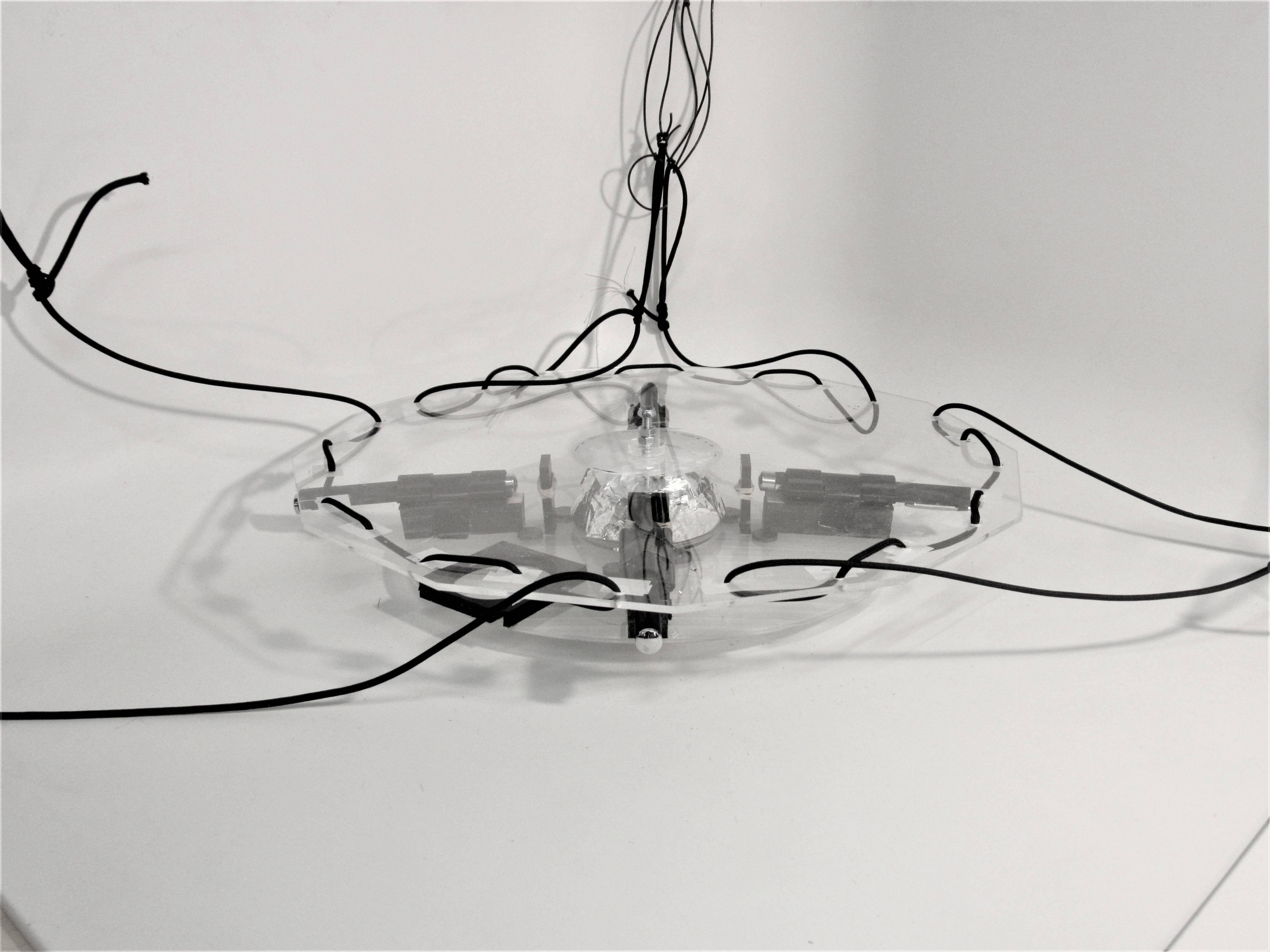 Suspended clear acrylic sculpture with central foil cone, laser mounts, and trailing black wires.