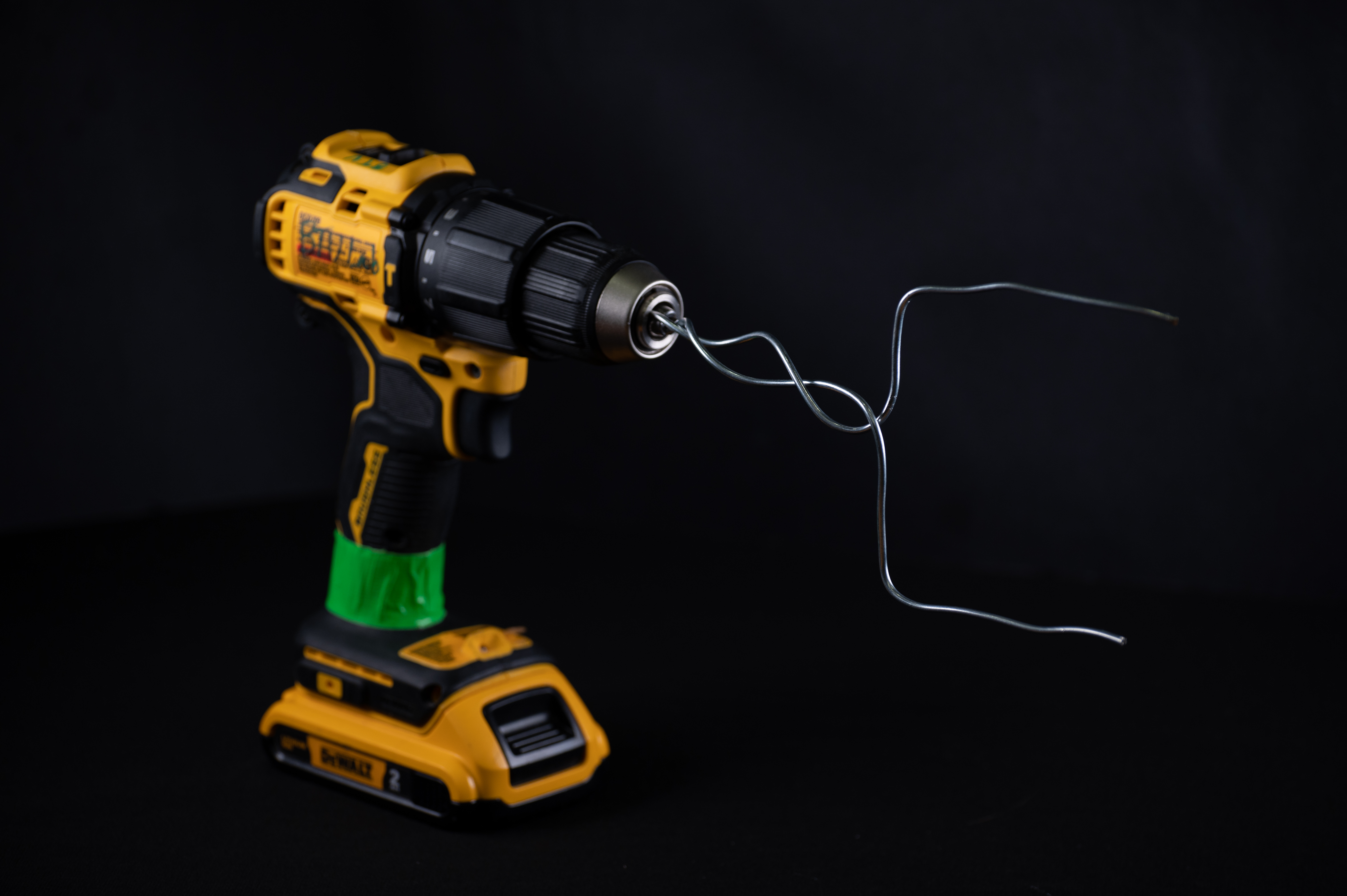A yellow DeWalt power drill twisting a thin metal wire tool against a black background.