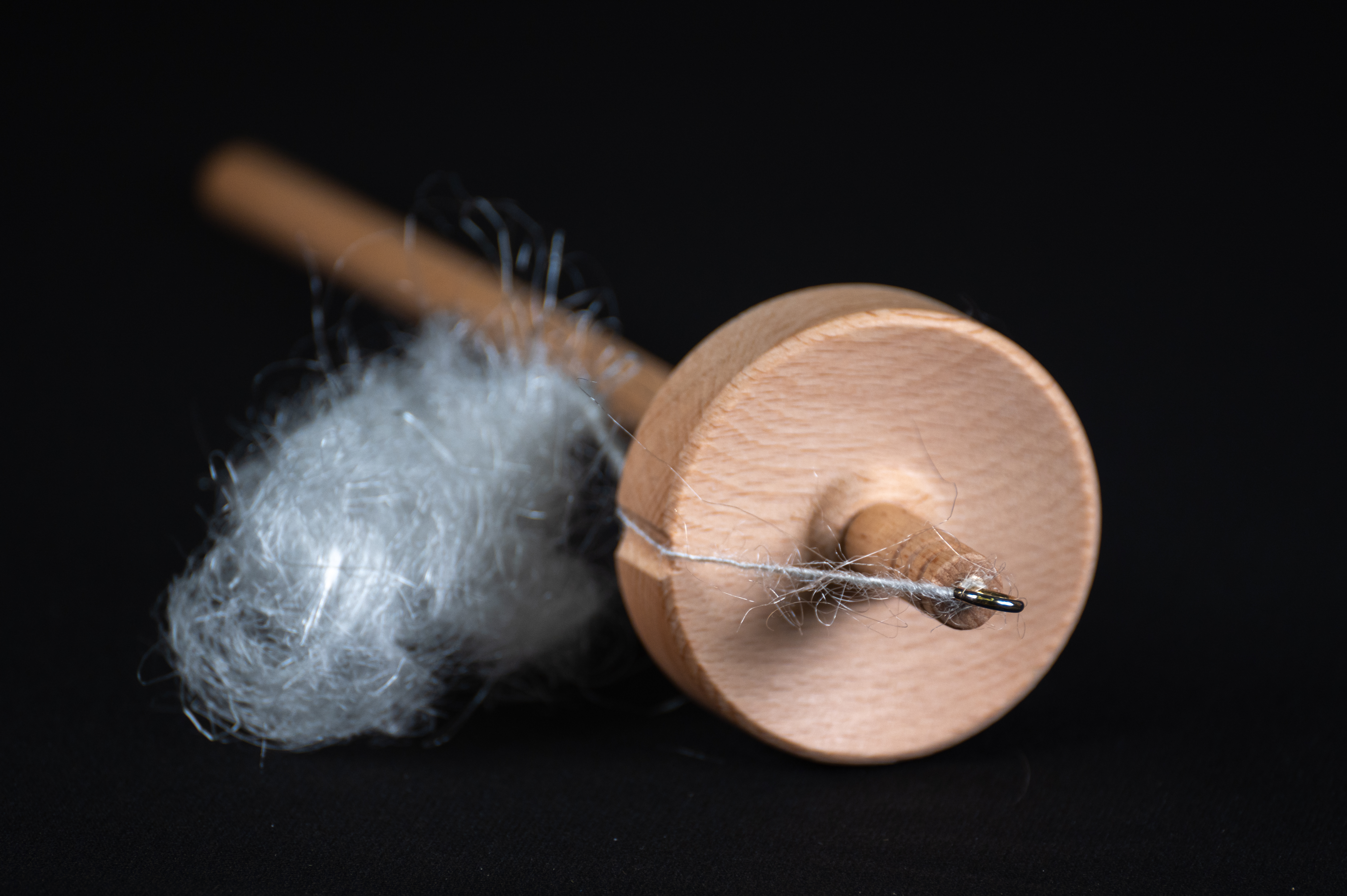 A wooden drop spindle winding white plastic fiber into yarn beside a fluff of roving.