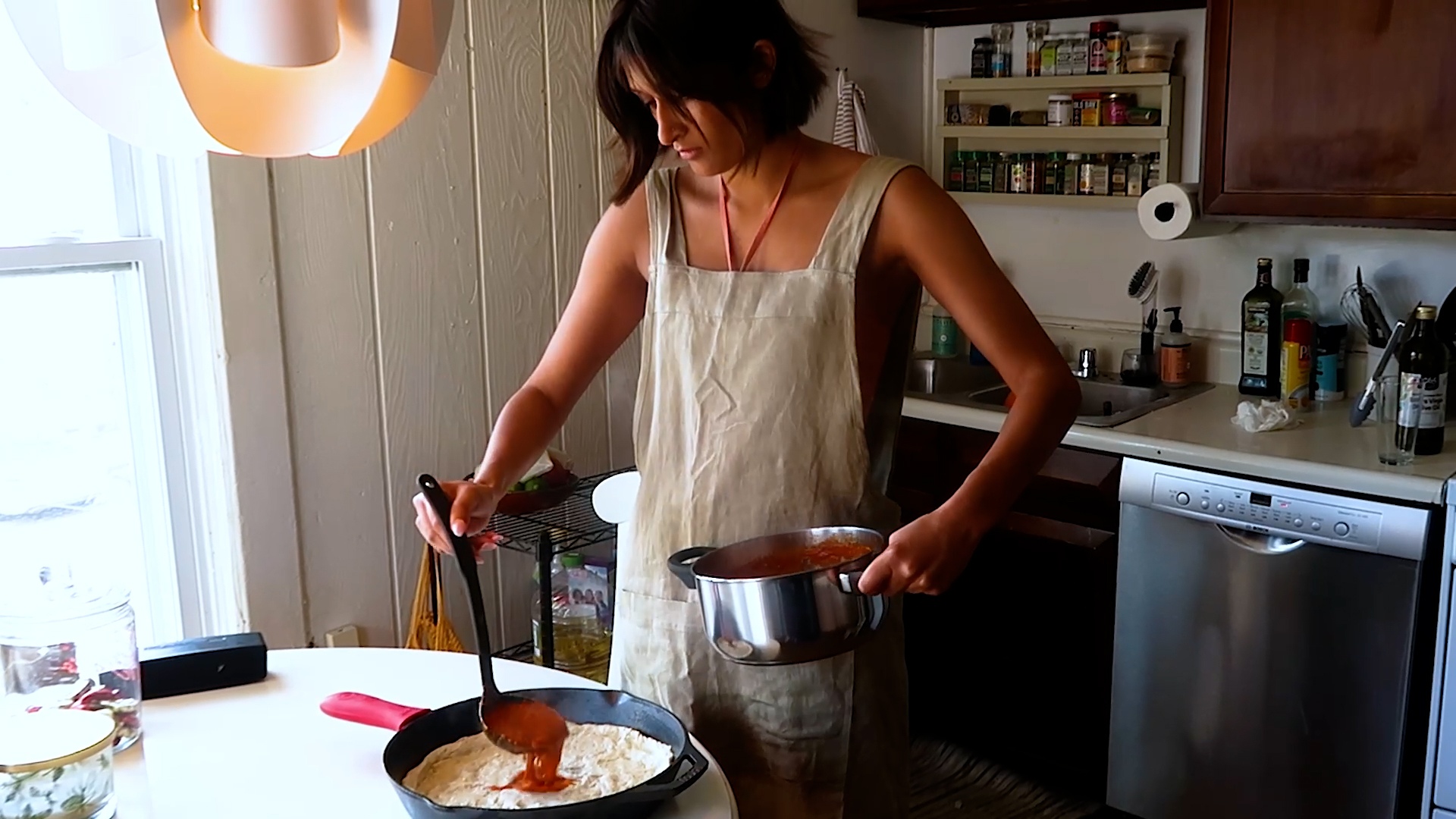 A woman in an apron ladles red sauce over batter in a skillet in a home kitchen.