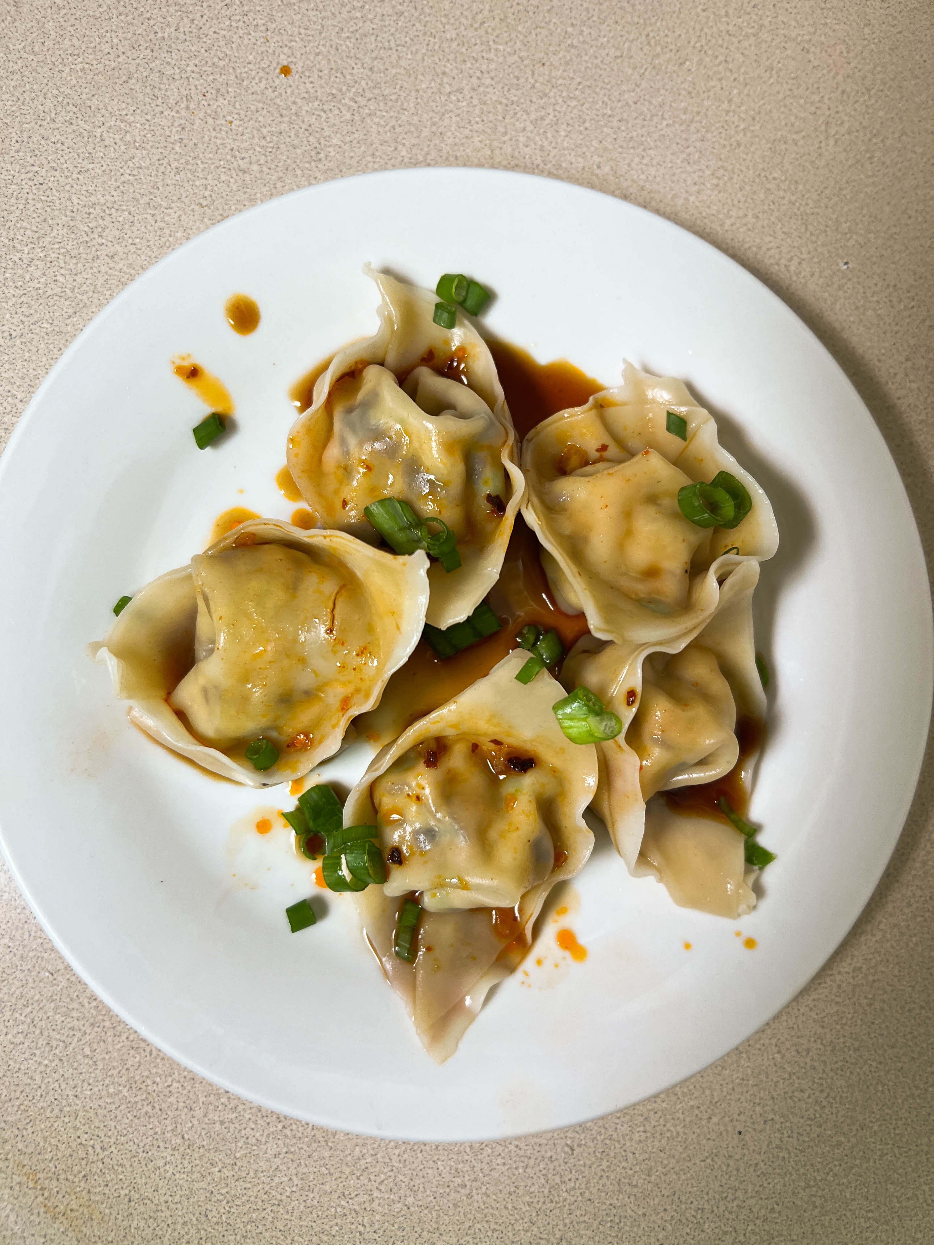 A white plate of steamed dumplings drizzled with chili oil and topped with green onions.