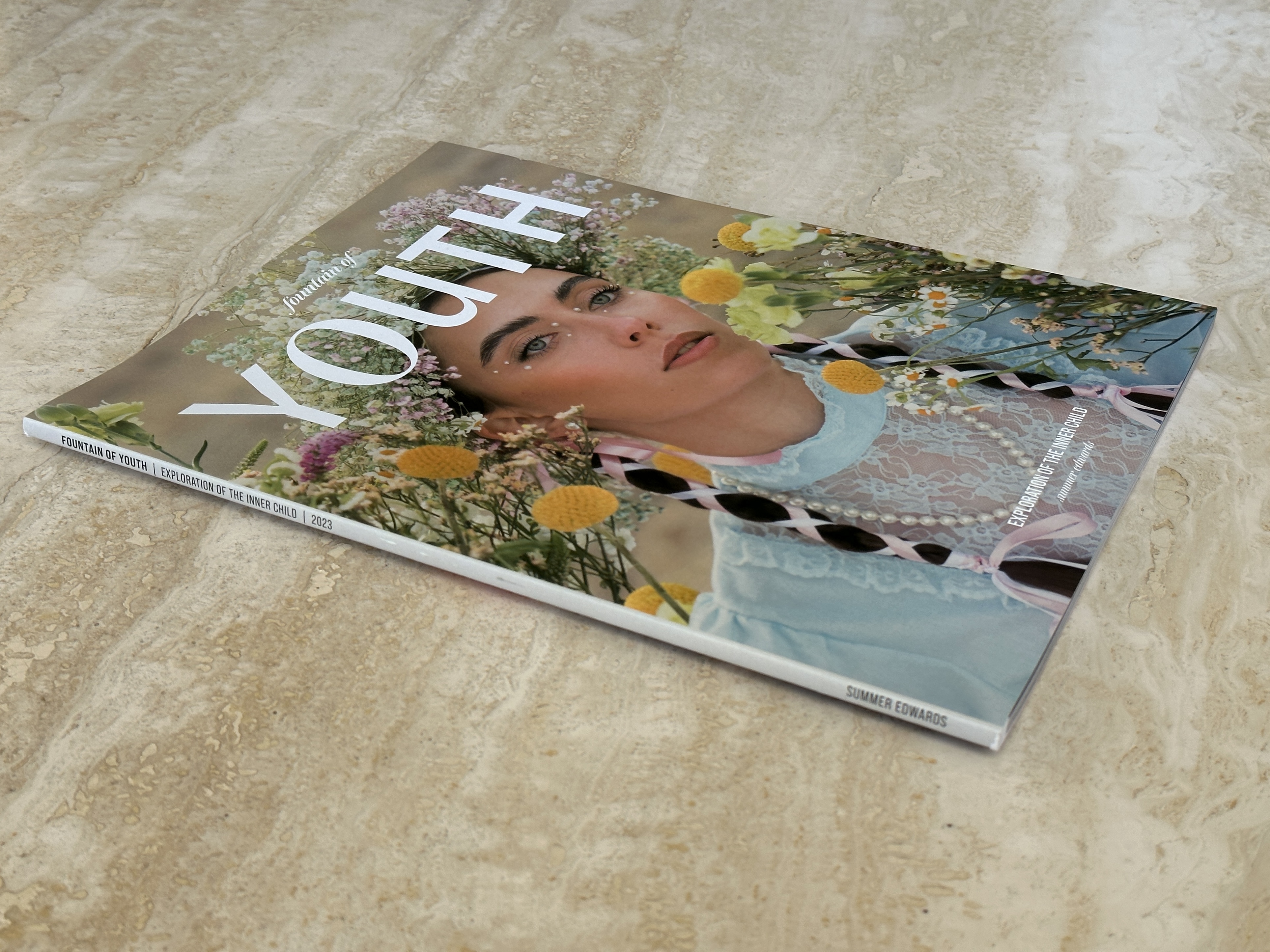 Printed Fountain of Youth magazine angled on stone, cover showing a flower-crowned model.