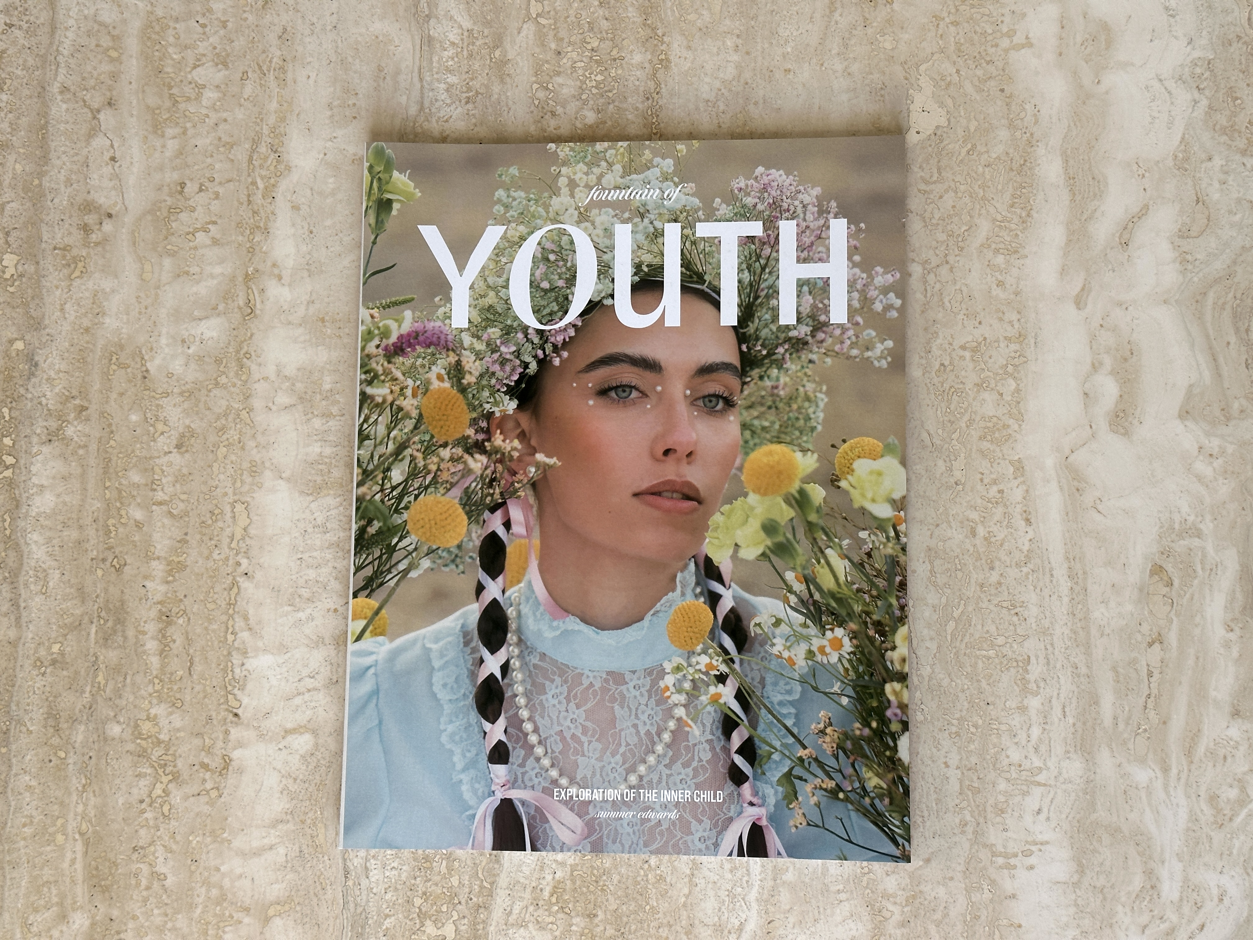 Fountain of Youth magazine flat on stone; cover model wears wildflowers, lace, and pearl-braided hair.