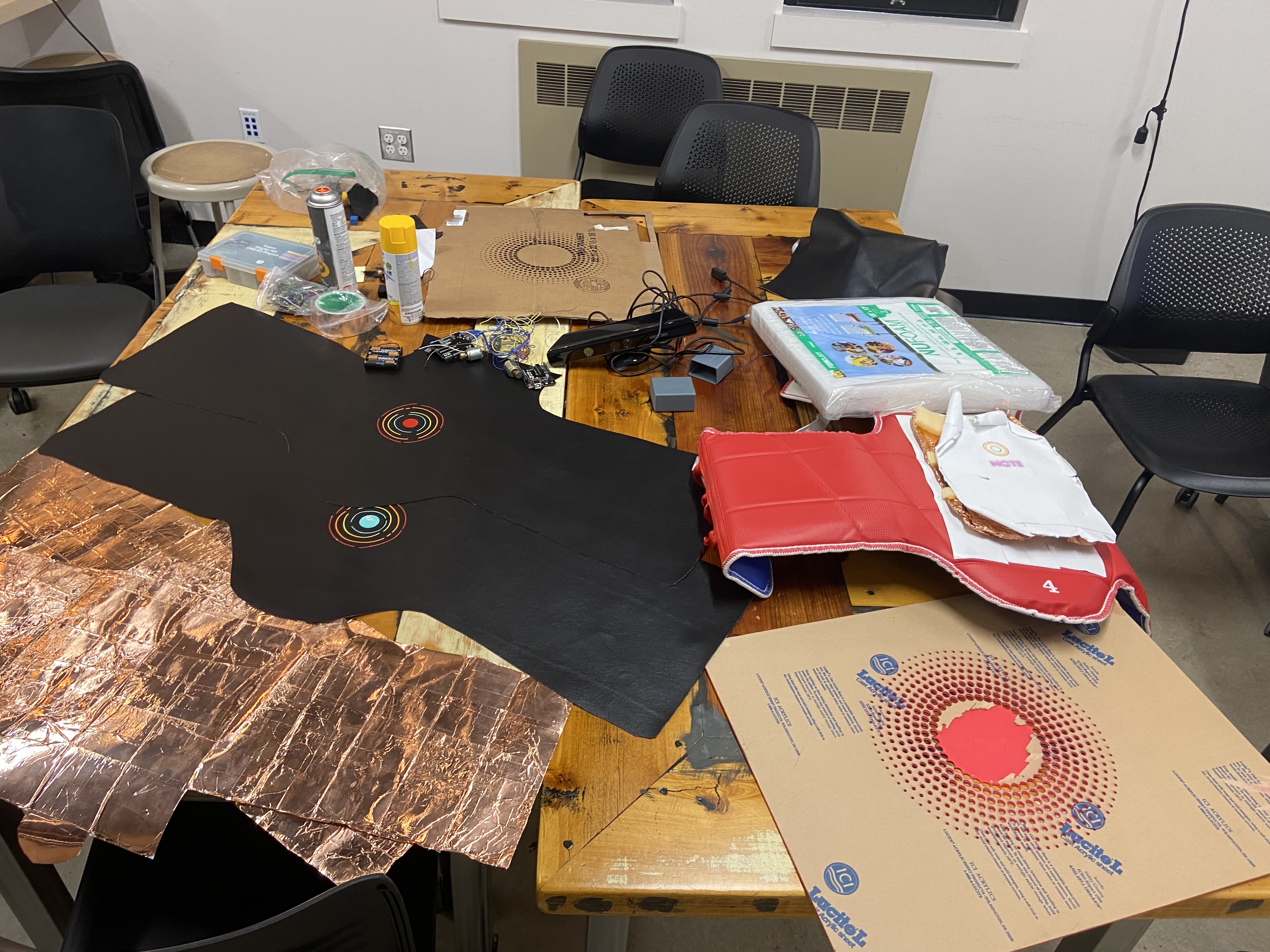 Workshop table strewn with black vest panels, copper sheets, wiring, and a red taekwondo chest guard.