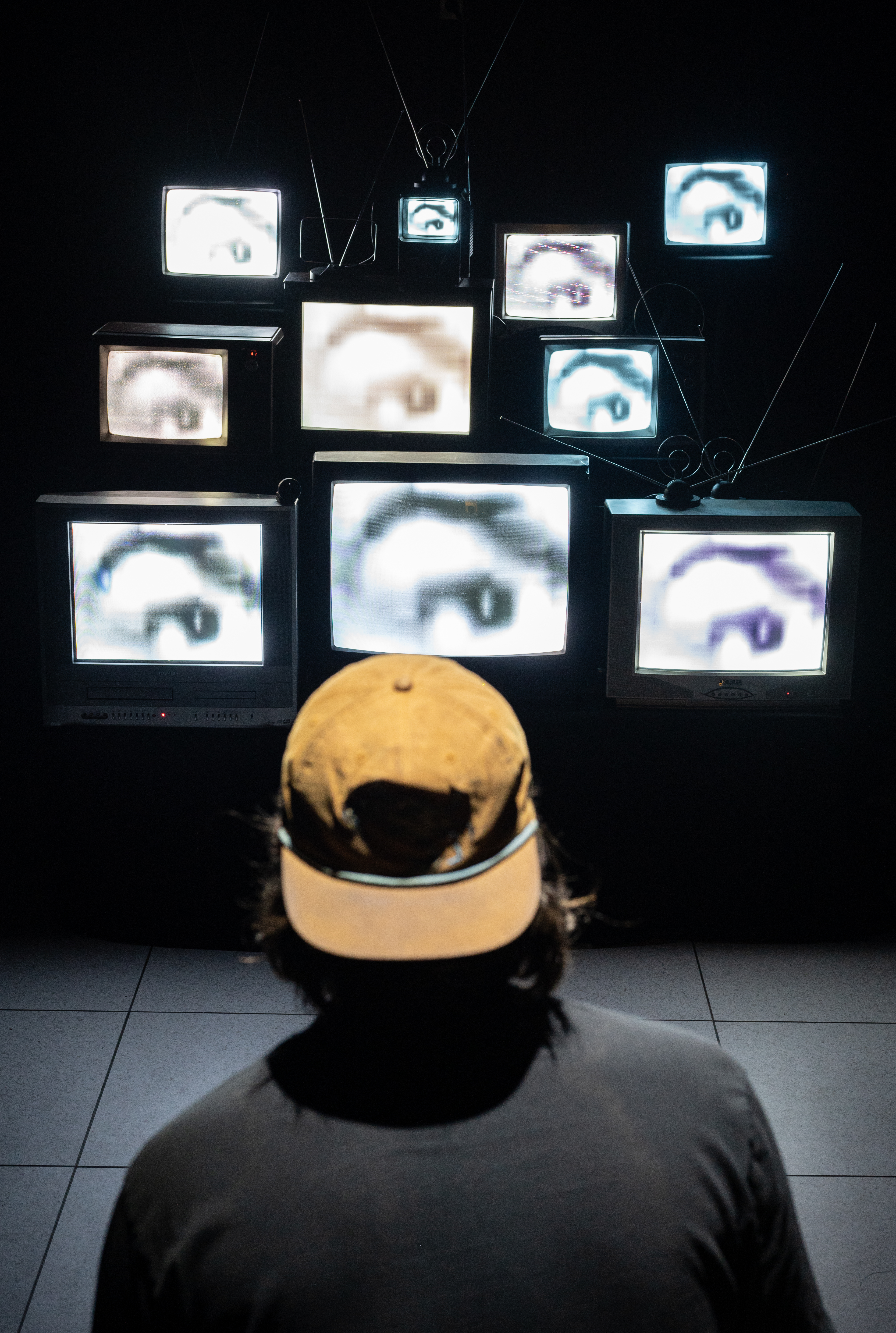 A capped viewer faces a dark wall of stacked CRT TVs, each showing a close-up of an eye.