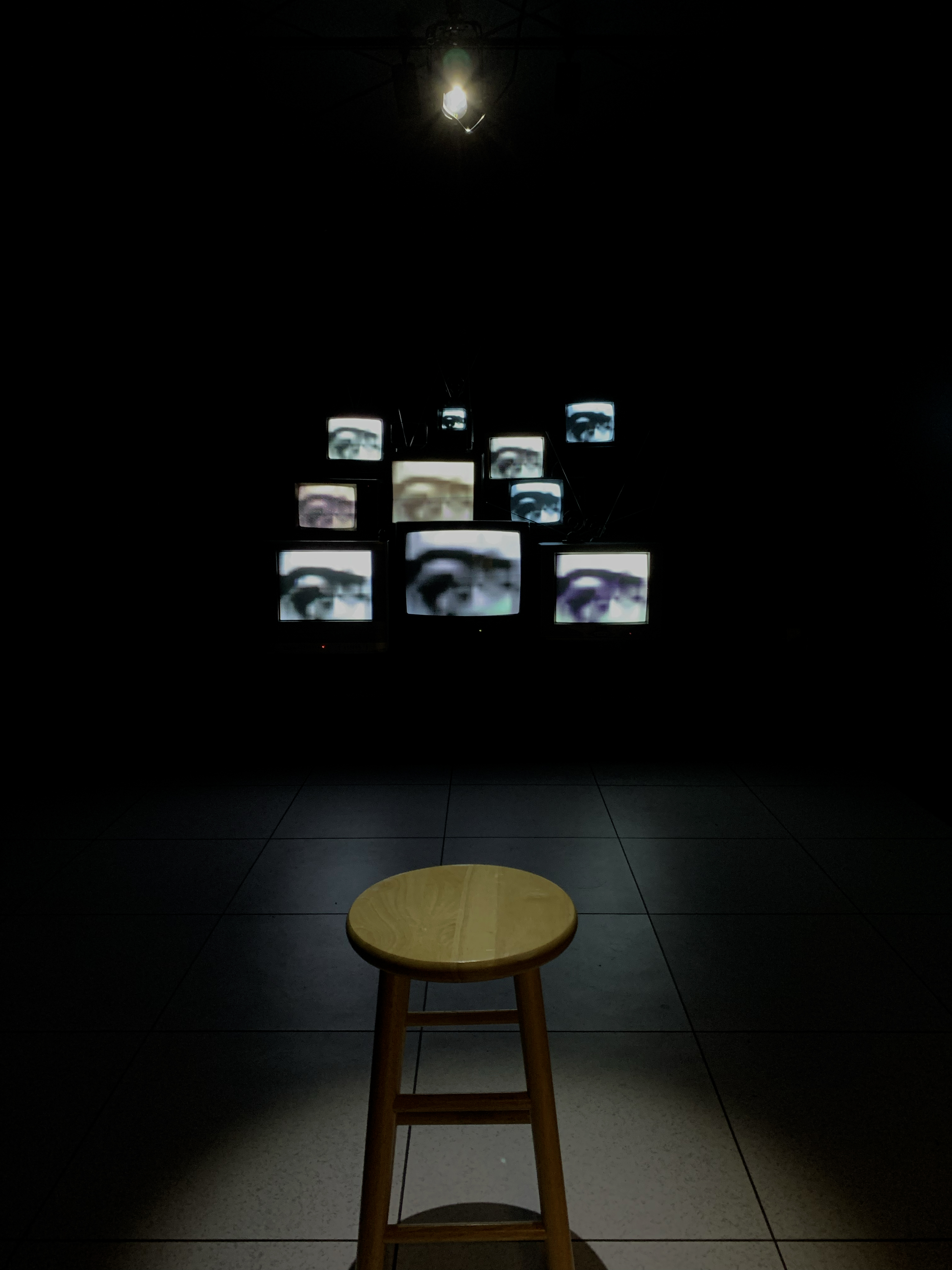 A lone wooden stool spotlit before a distant cluster of CRT TVs glowing with eye imagery in a dark room.