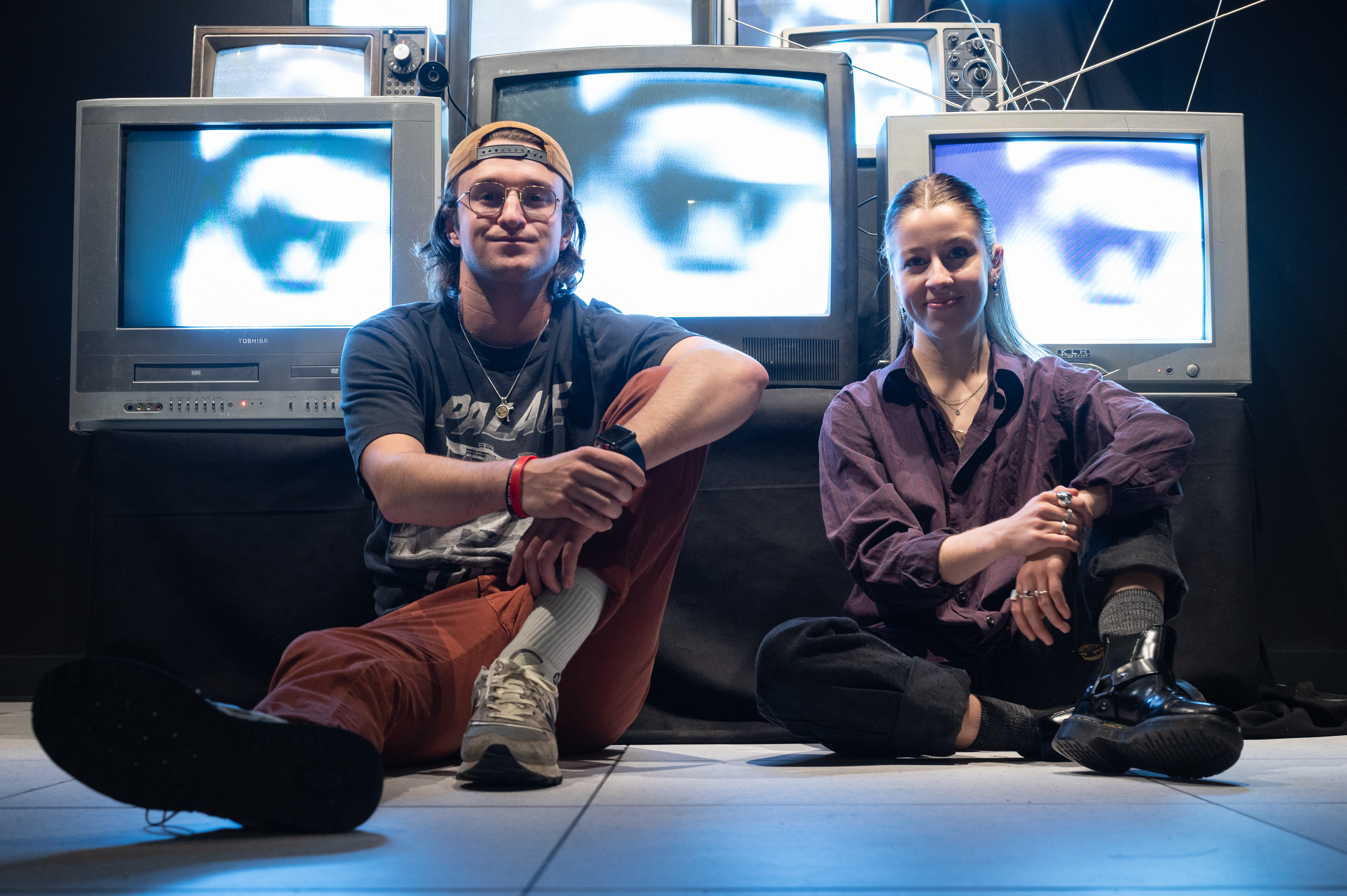 Two smiling creators sit on the floor before their wall of CRT TVs displaying close-up eyes.