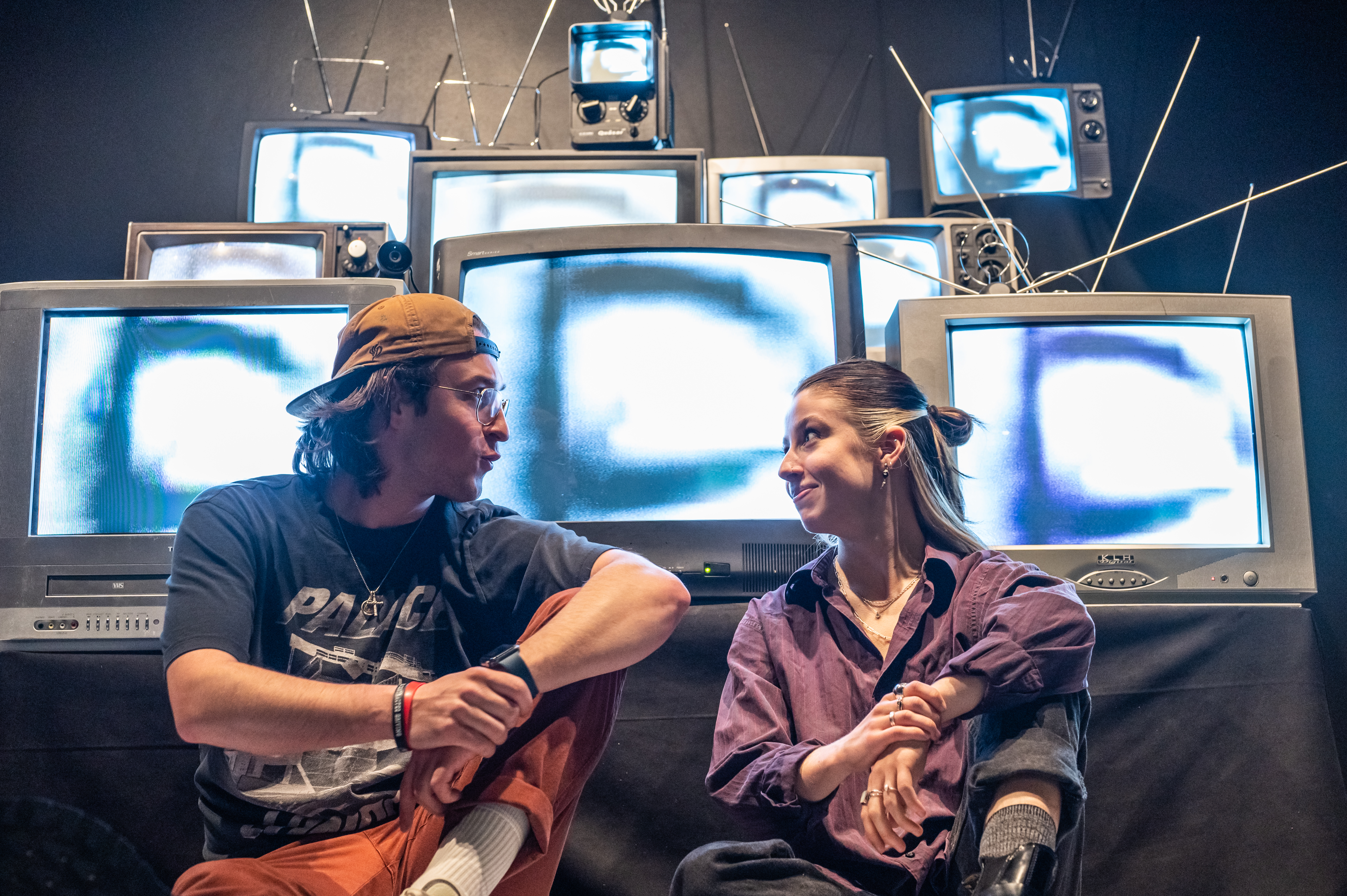 The two creators sit facing each other in front of their stacked wall of glowing vintage CRT TVs.
