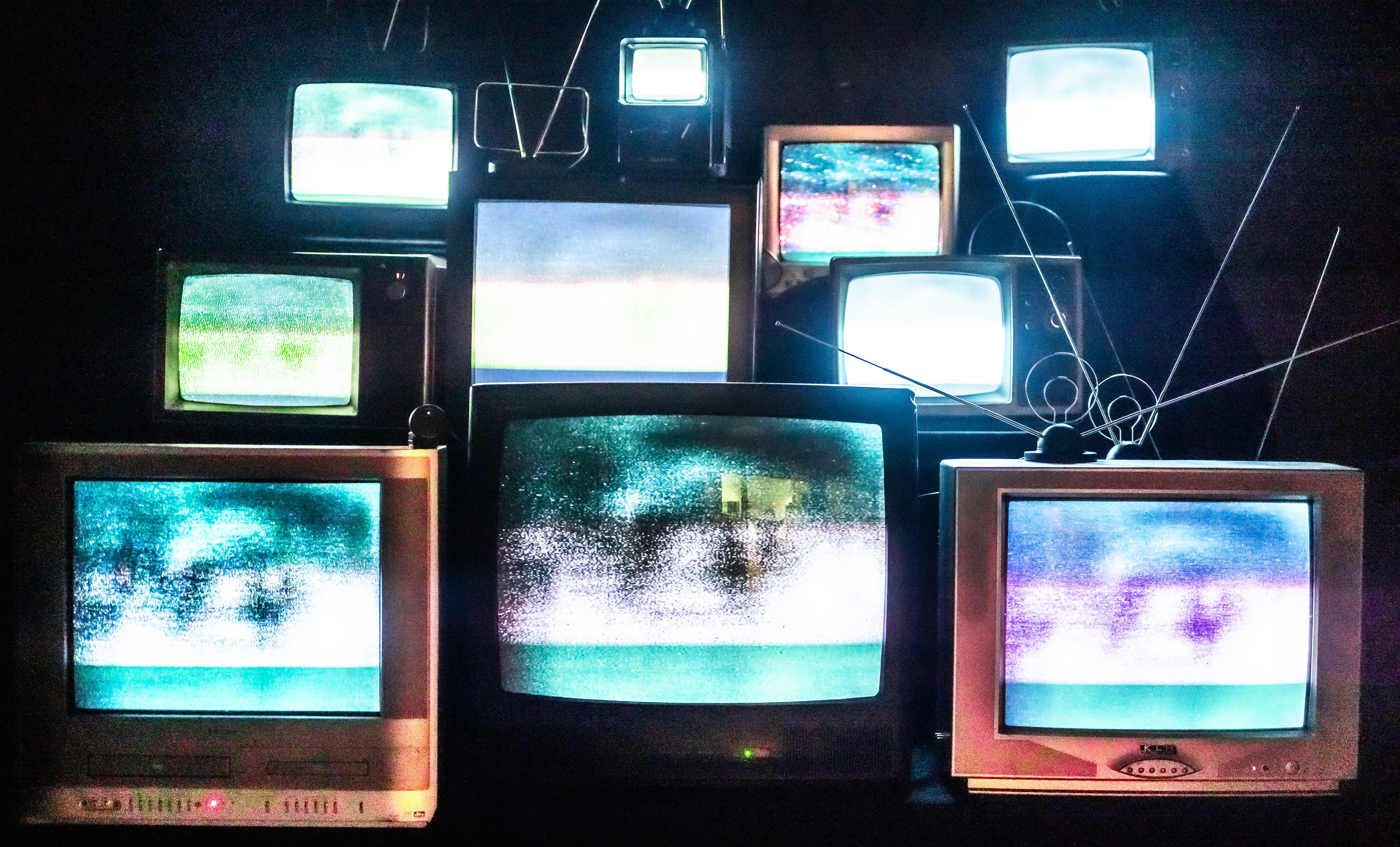 A staggered wall of vintage CRT televisions glowing with staticky teal, green, and purple imagery.