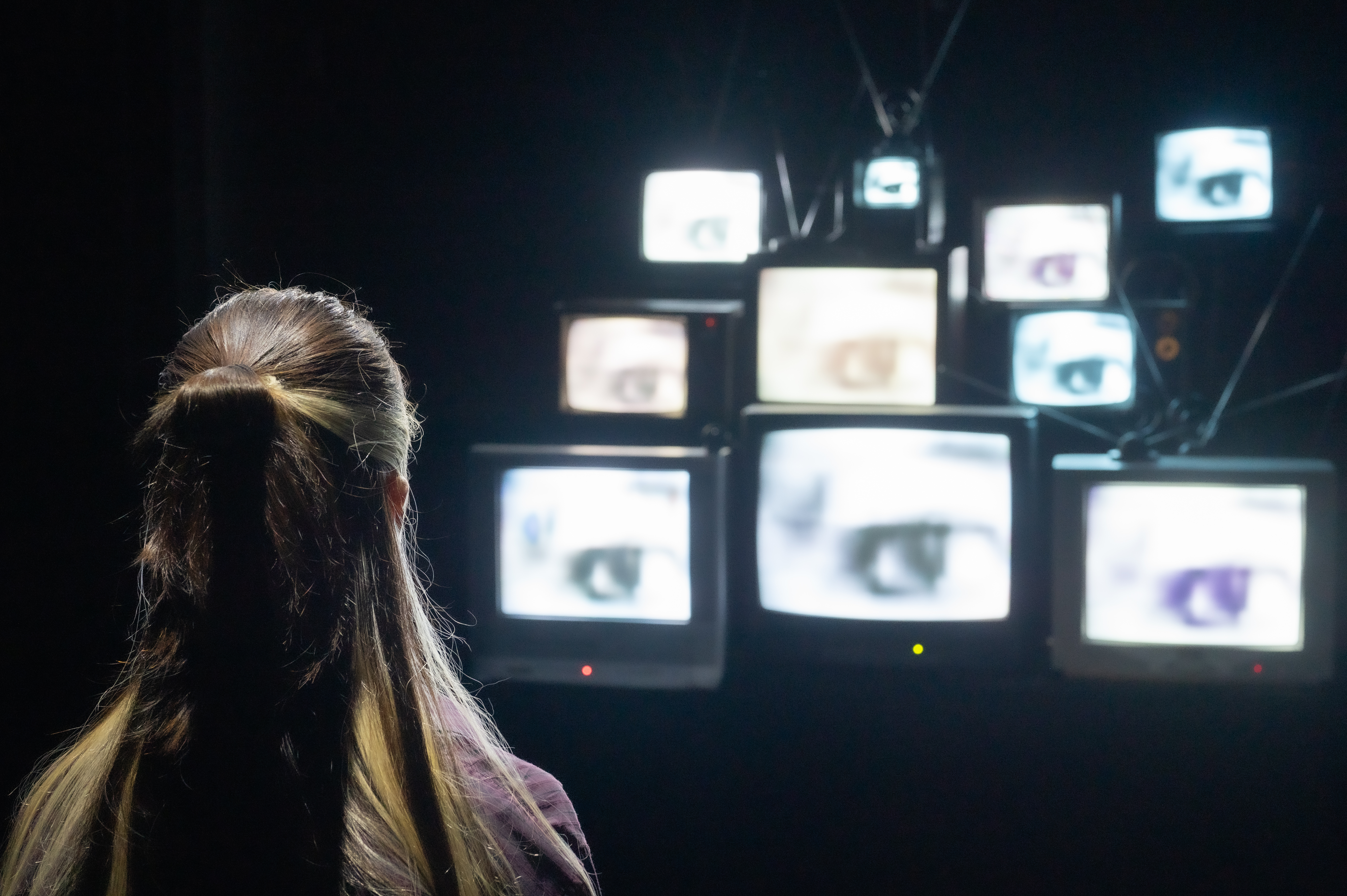 A long-haired viewer's silhouette faces a dim cluster of CRT TVs each showing a close-up eye.