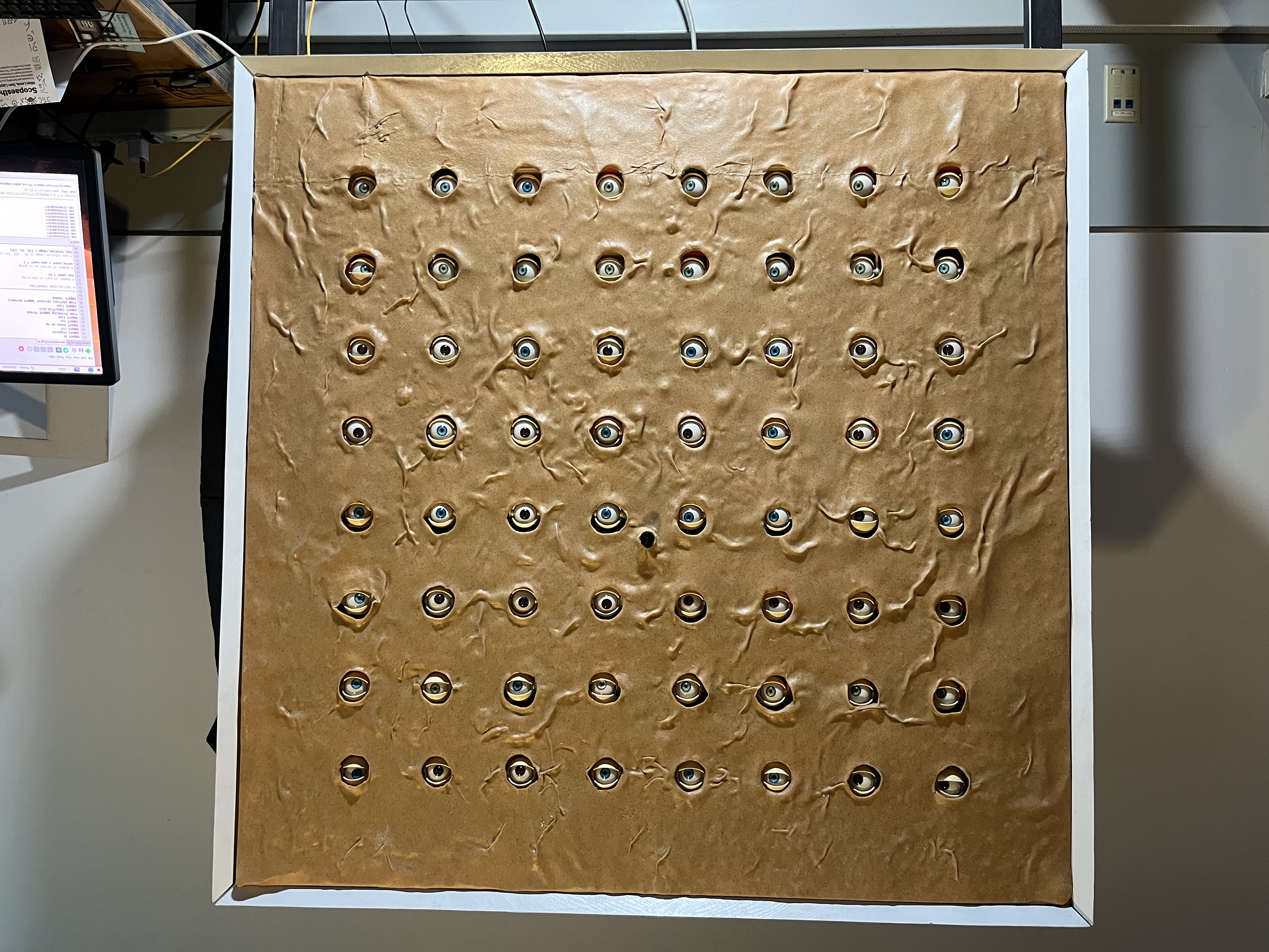 A square panel covered in textured brown skin with a grid of dozens of realistic animatronic eyes.
