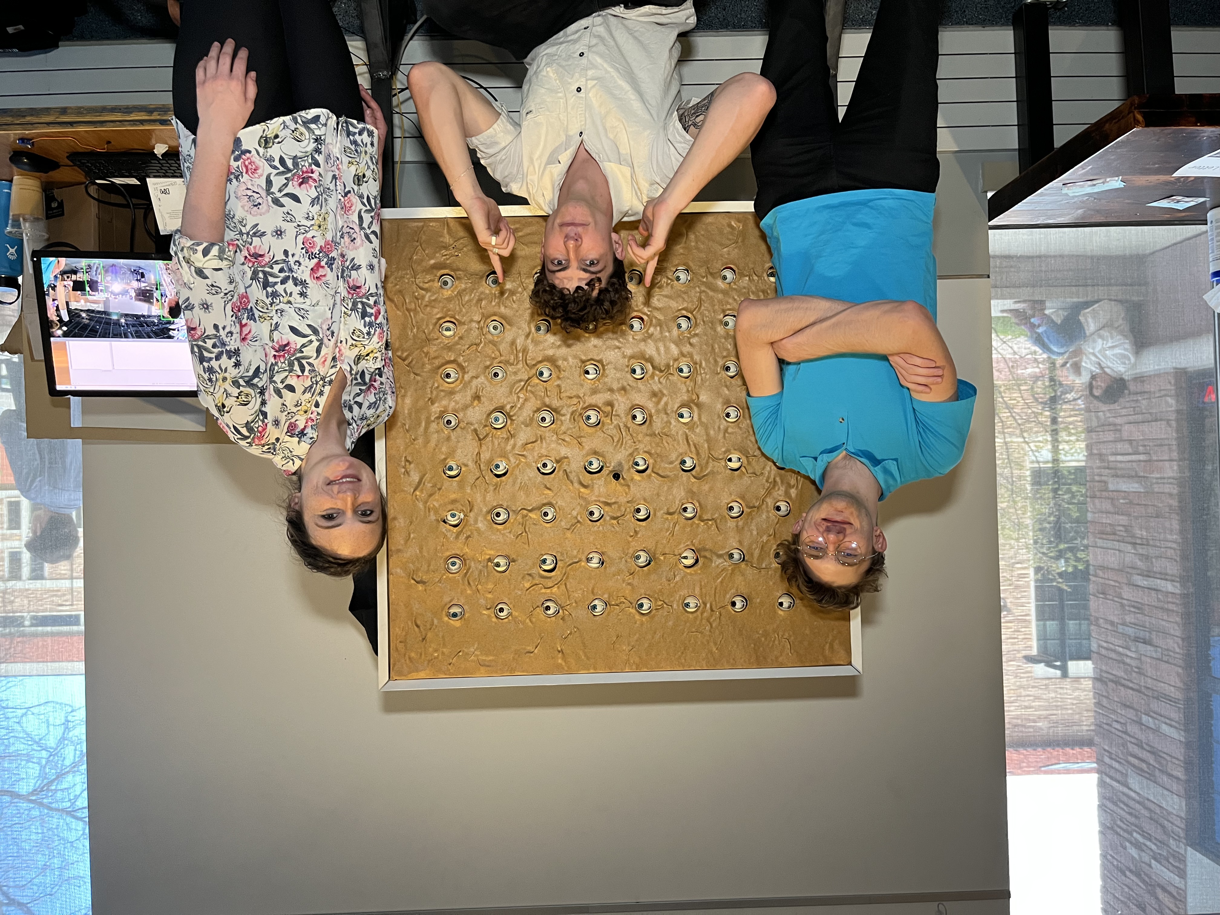 Three students lean over a panel of dozens of embedded animatronic eyes in a studio.