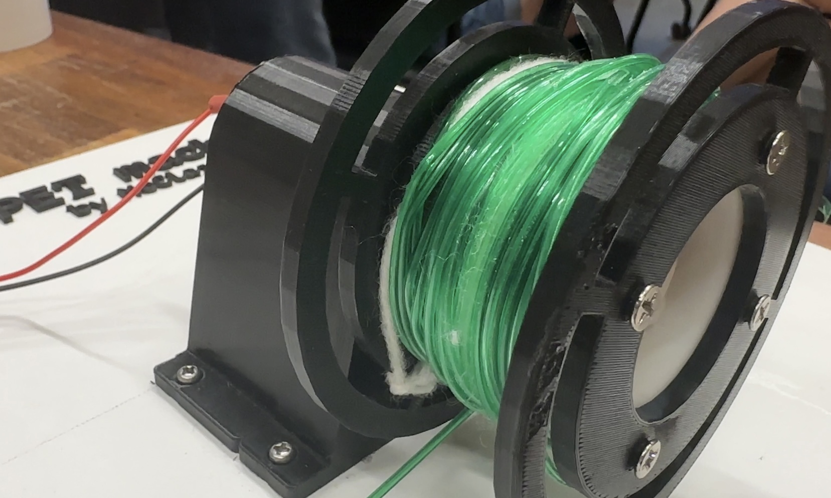 A black 3D-printed spool winding bright green recycled plastic filament.