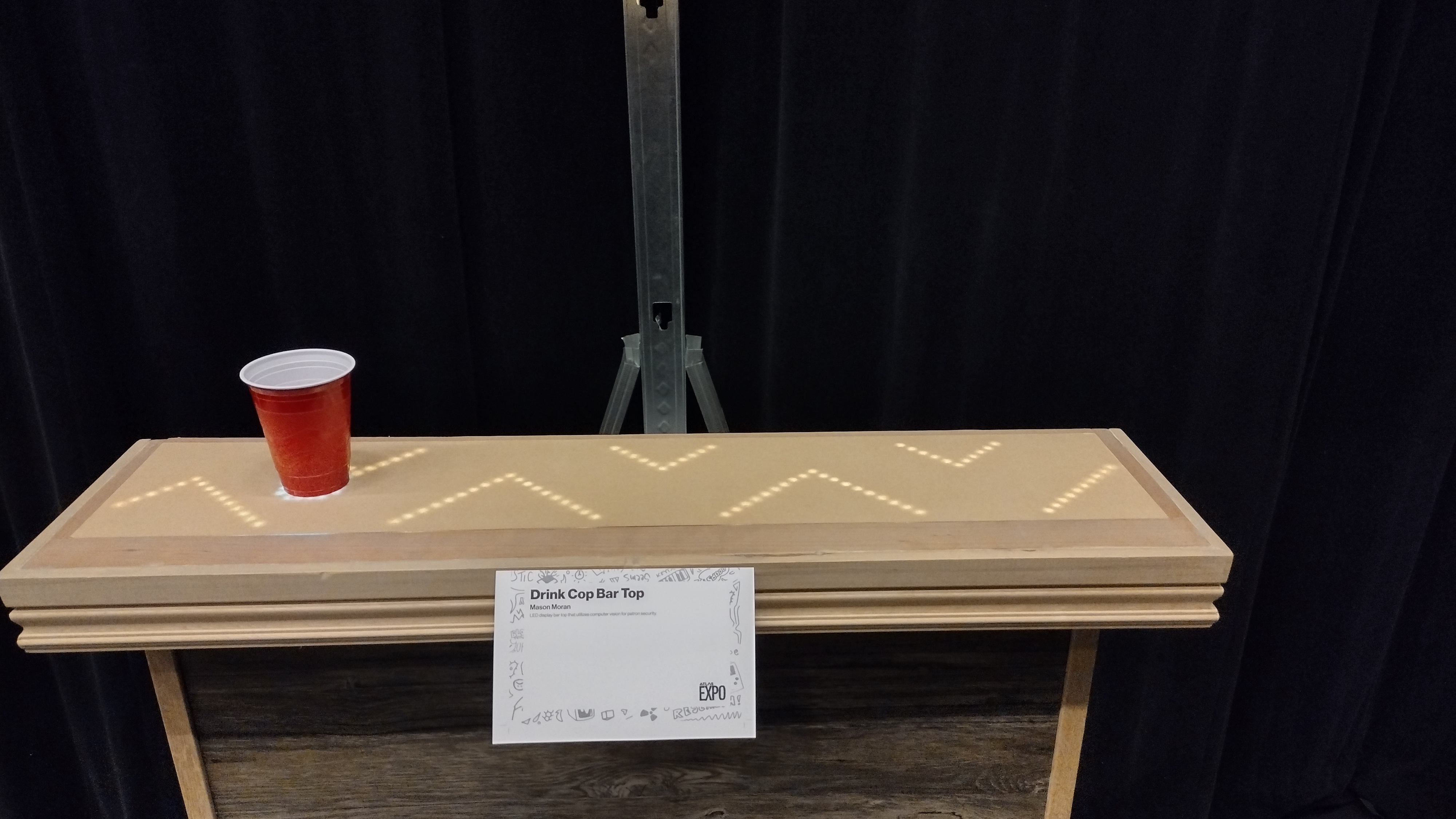 A wooden bar top with an embedded LED zigzag pattern, a red cup, and a project placard.