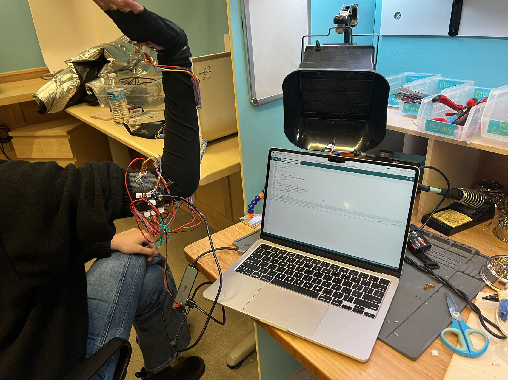 A student wearing a wired sensor sleeve on their arm at a workbench with a laptop.