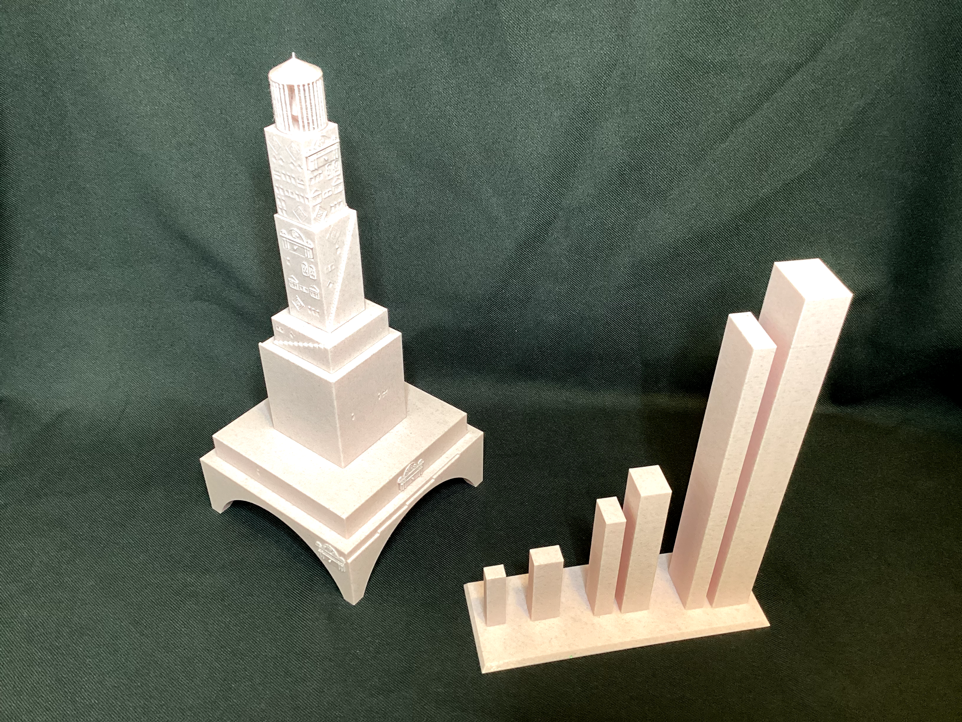 Two white 3D-printed data sculptures, a tiered tower and a rising bar chart, on green fabric.
