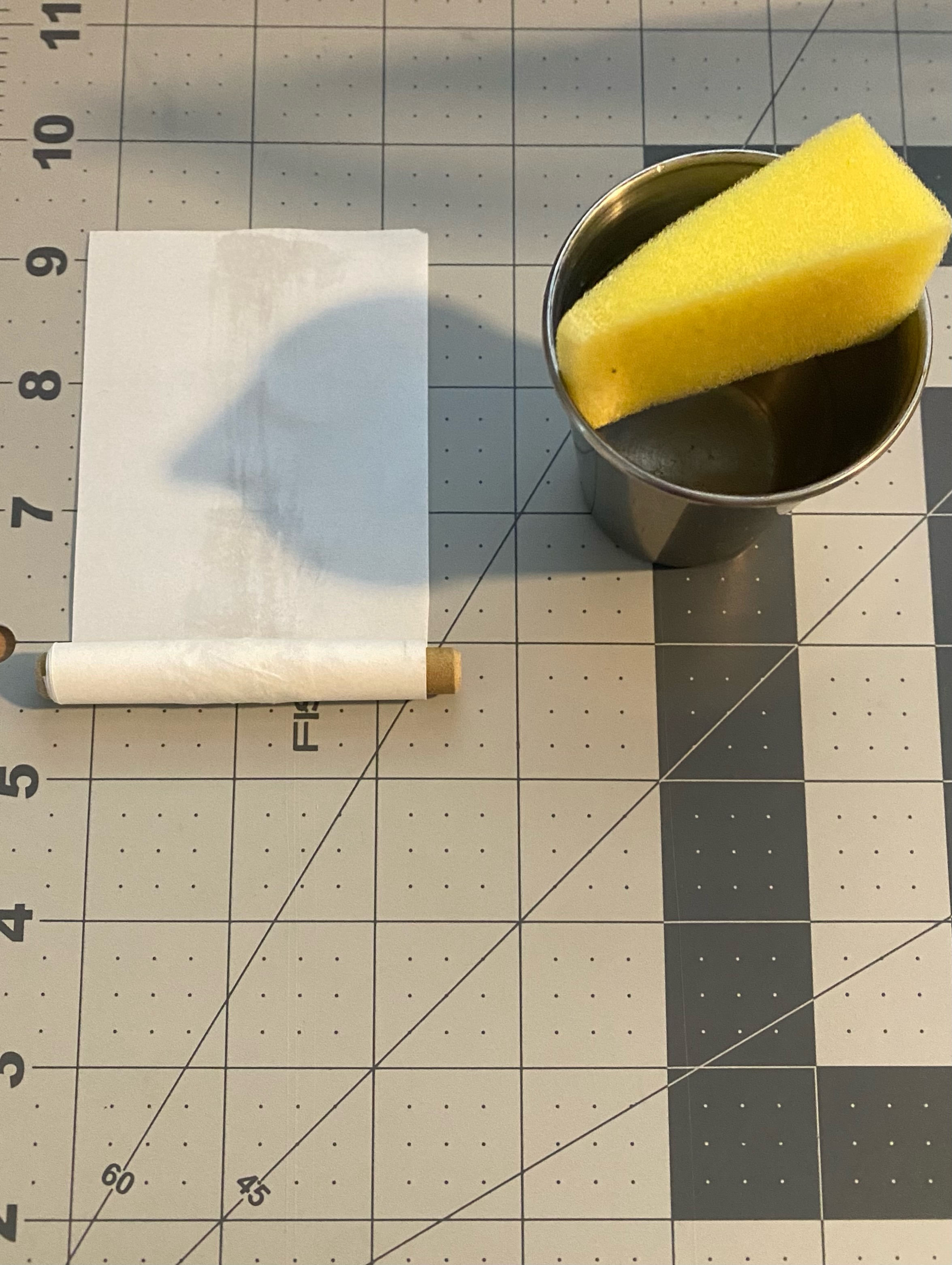 A paper sheet partly rolled around a dowel beside a damp sponge in a metal cup, on a cutting mat.