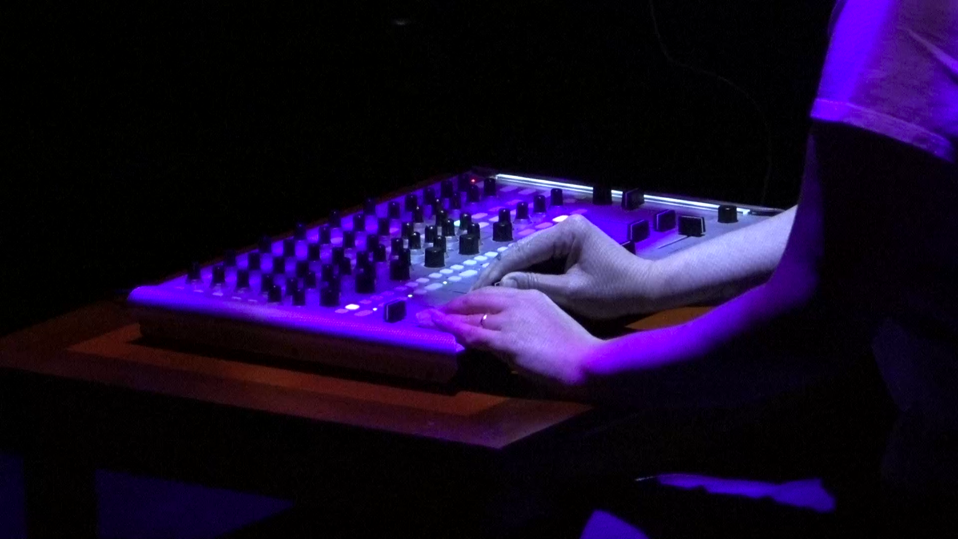 Hands play a knob-covered synthesizer lit with purple light in a dark theater.