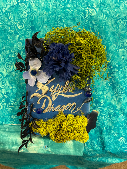 A decorated blue book titled Sapphire Dragon in gold, adorned with flowers, moss, and dark leaves.