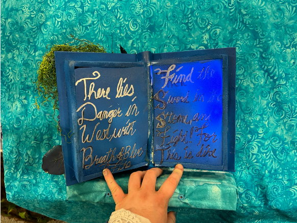 The open blue book with handwritten gold riddle verses, the right page glowing blue, a hand pointing.