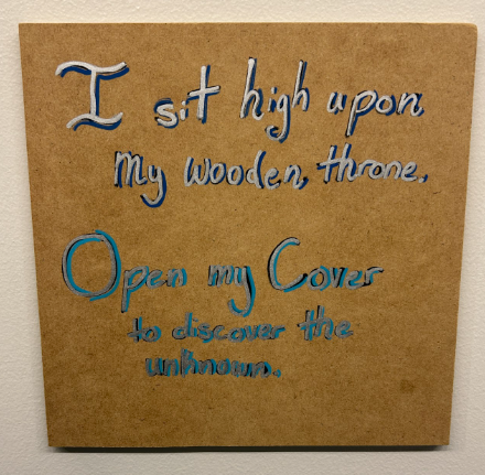 Wooden plaque with a blue rhyme: I sit high upon my wooden throne, open my cover to discover the unknown.