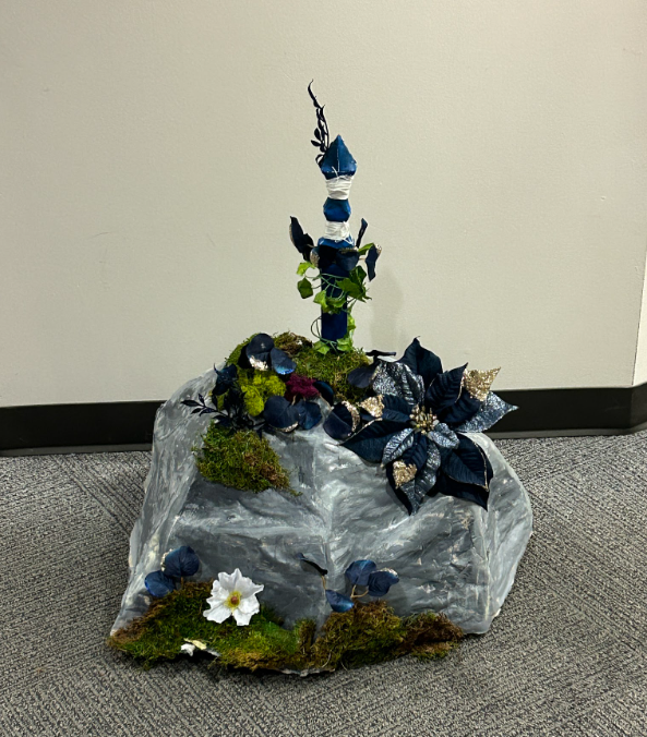 A sword-in-the-stone prop: a blue hilt rising from a moss-covered grey rock with blue flowers.