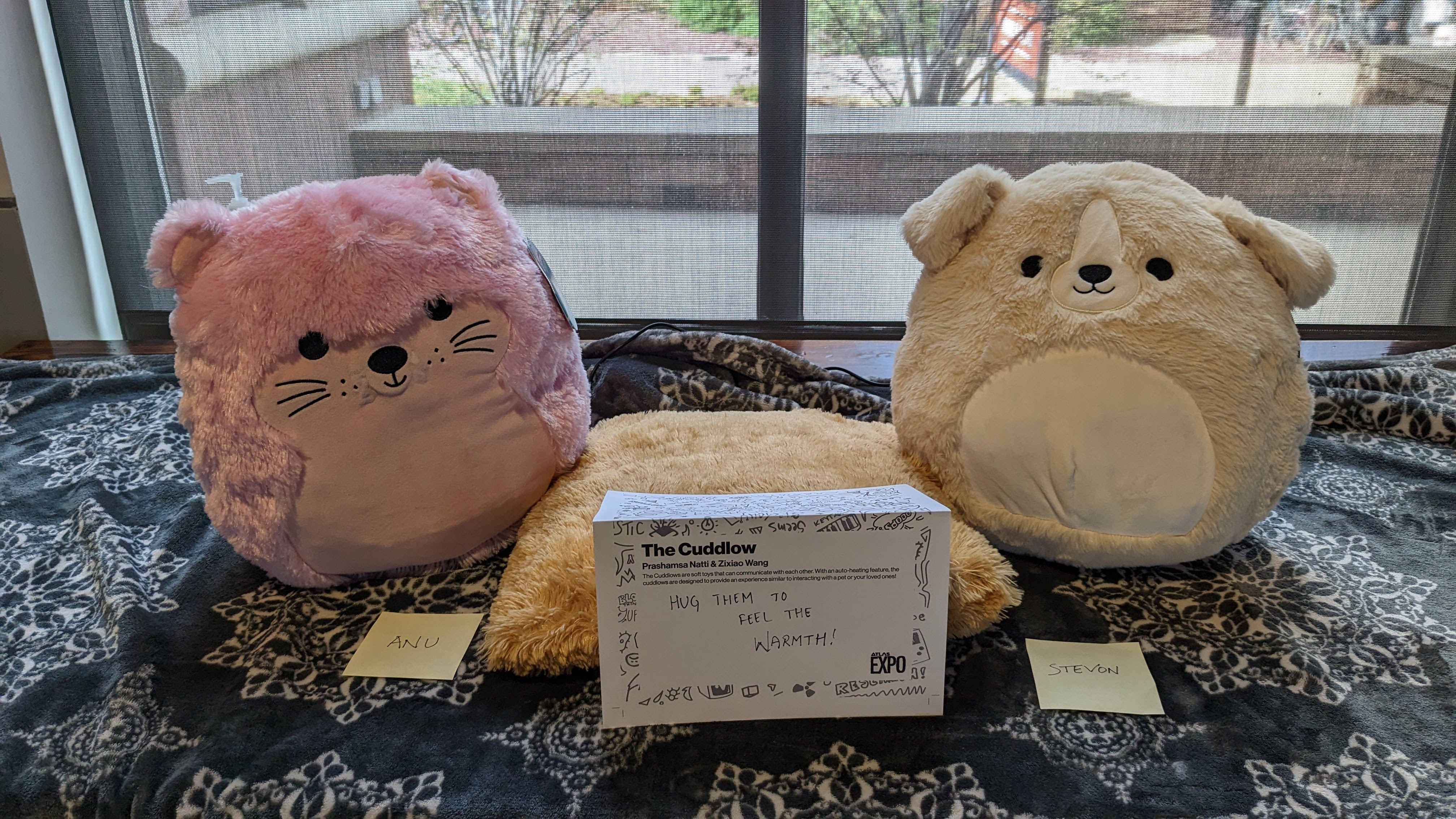 Two round plush toys, a pink cat and tan bear, with a placard reading Hug them to feel the warmth.