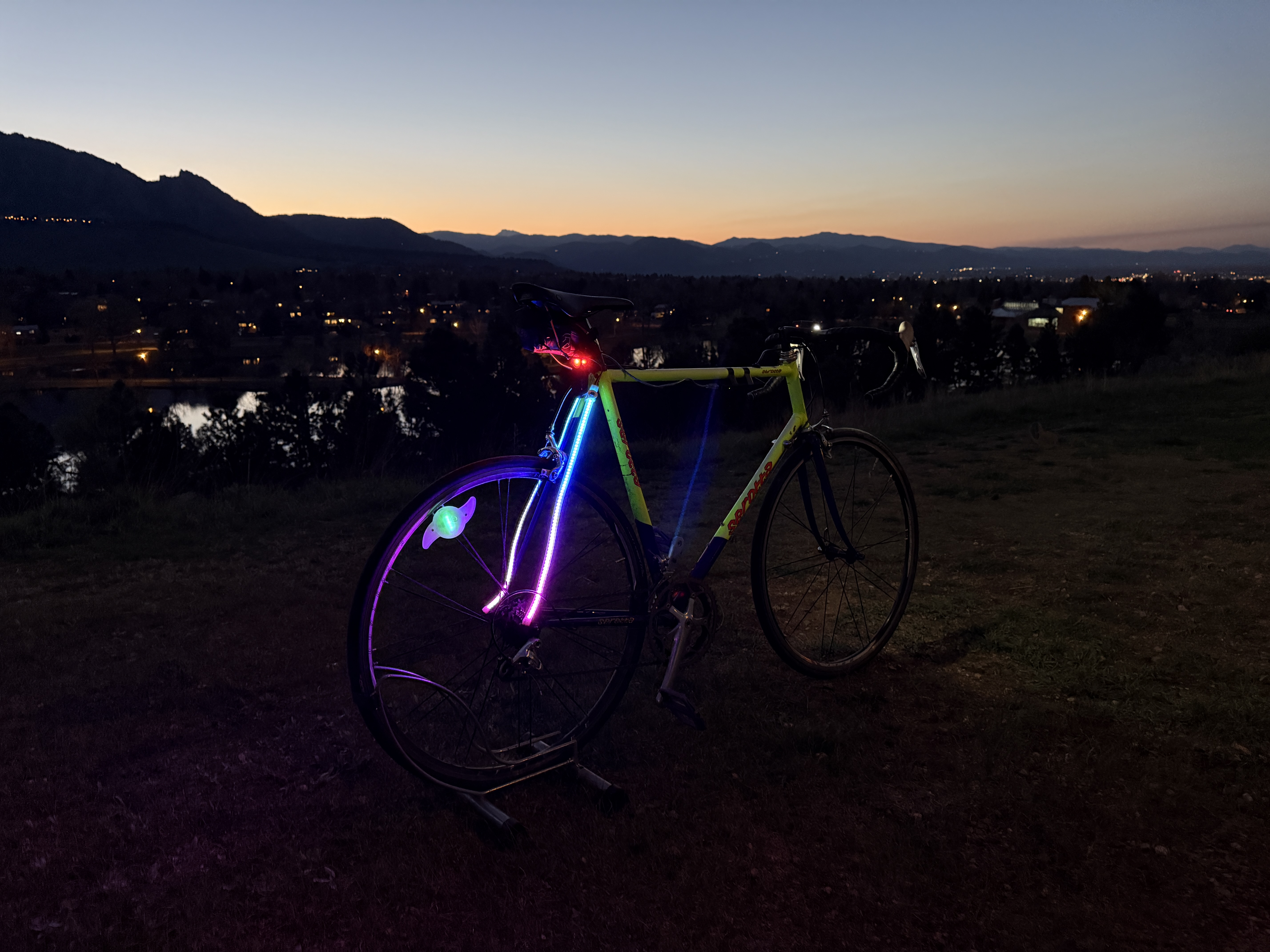 Road bike on a stand at dusk, frame and spokes lit with rainbow LED strips, mountains behind.