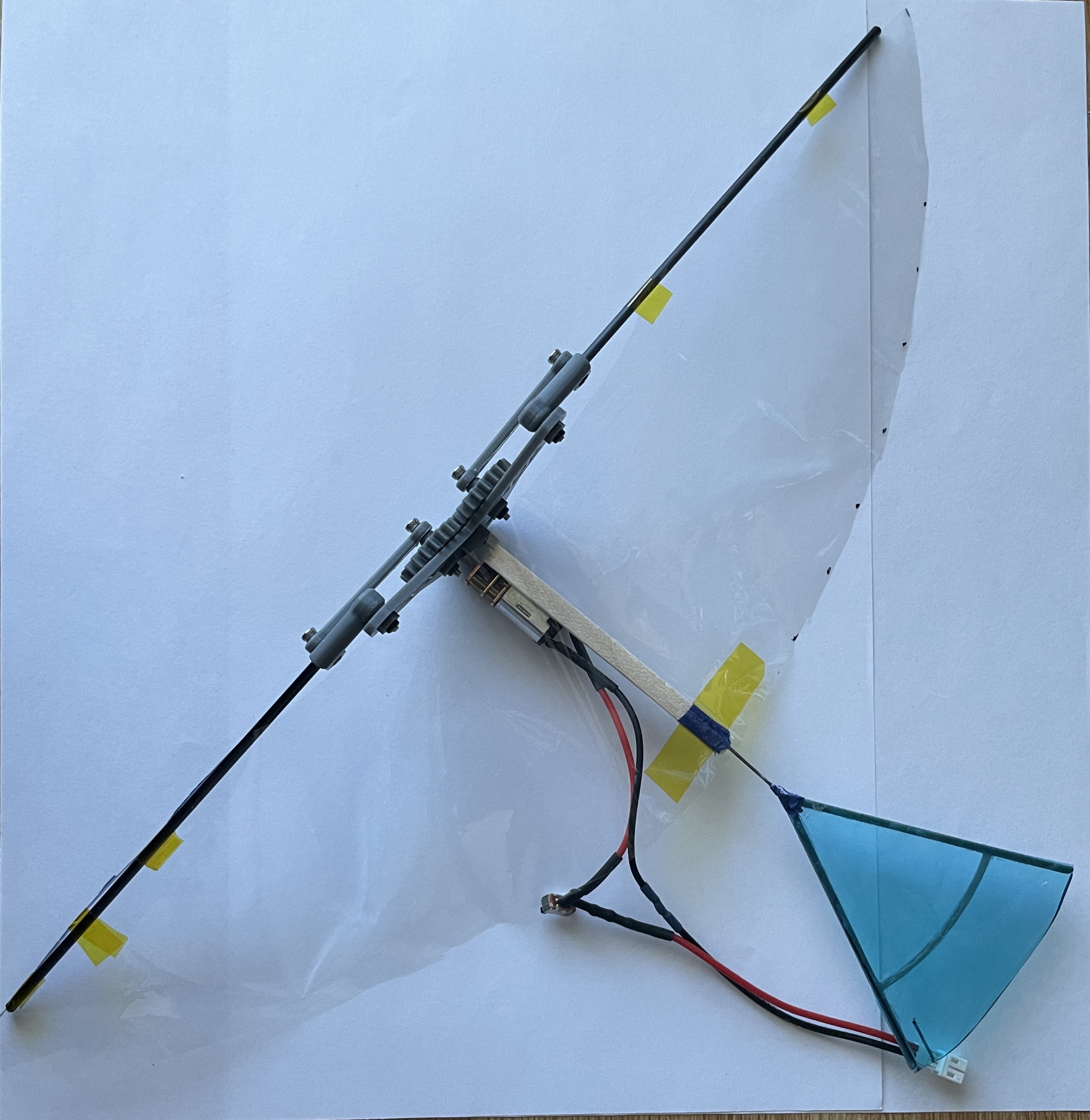 A prototype ornithopter with a gear mechanism, clear film wings, and a blue tail, taped on white paper.