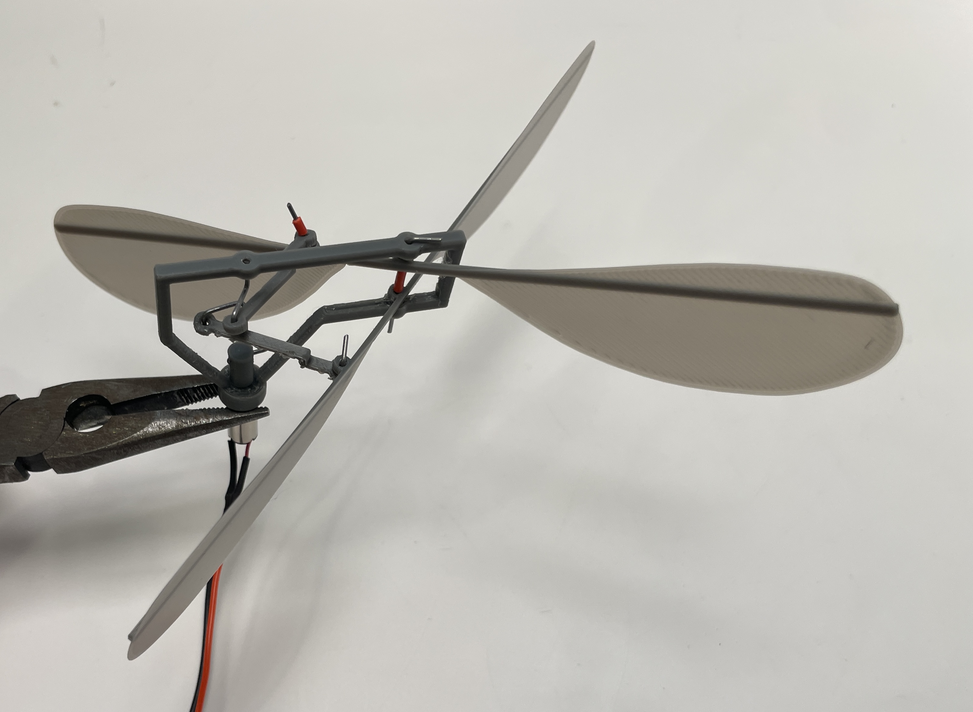 Close-up of an ornithopter's grey 3D-printed flapping mechanism and membrane wings, held by pliers.
