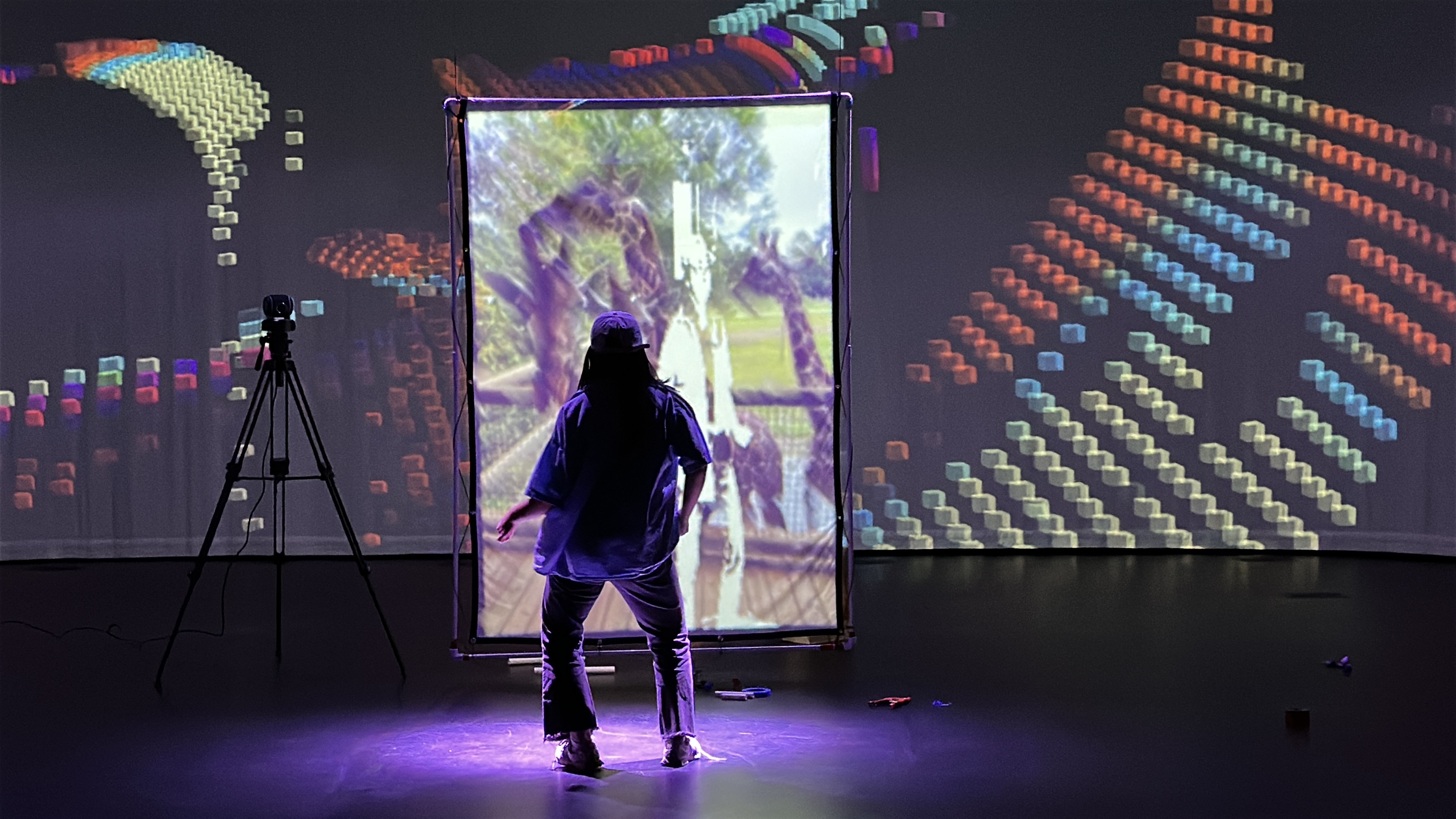 A person stands before a translucent projection screen in a dark room filled with colorful pixel-block projections.