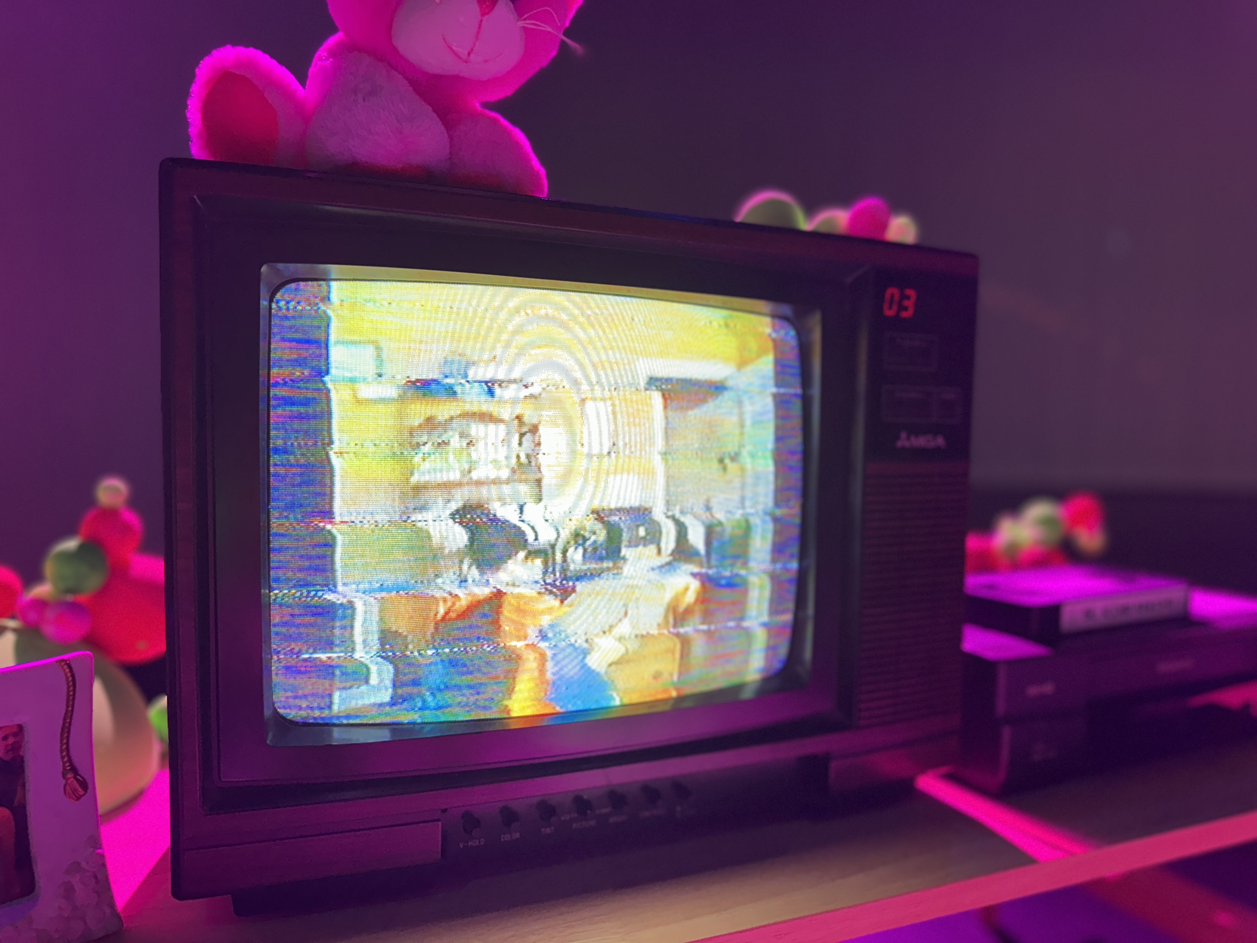 A vintage CRT TV showing glitchy static footage, topped with a pink stuffed animal, in purple light.